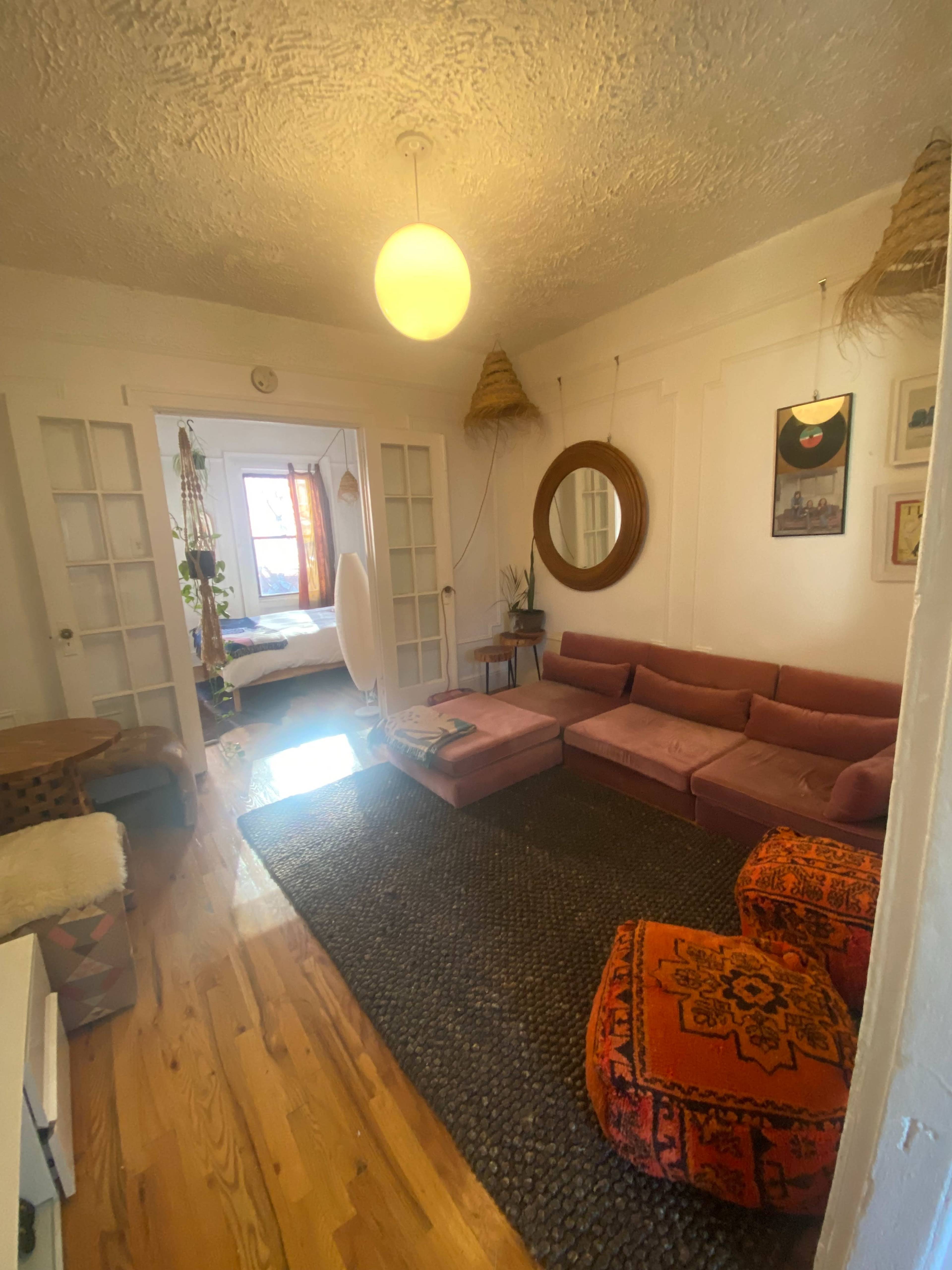A cozy living room features a pink sectional couch, a round mirror, and decorative items, with a glimpse of a bedroom visible in the background through an open door.