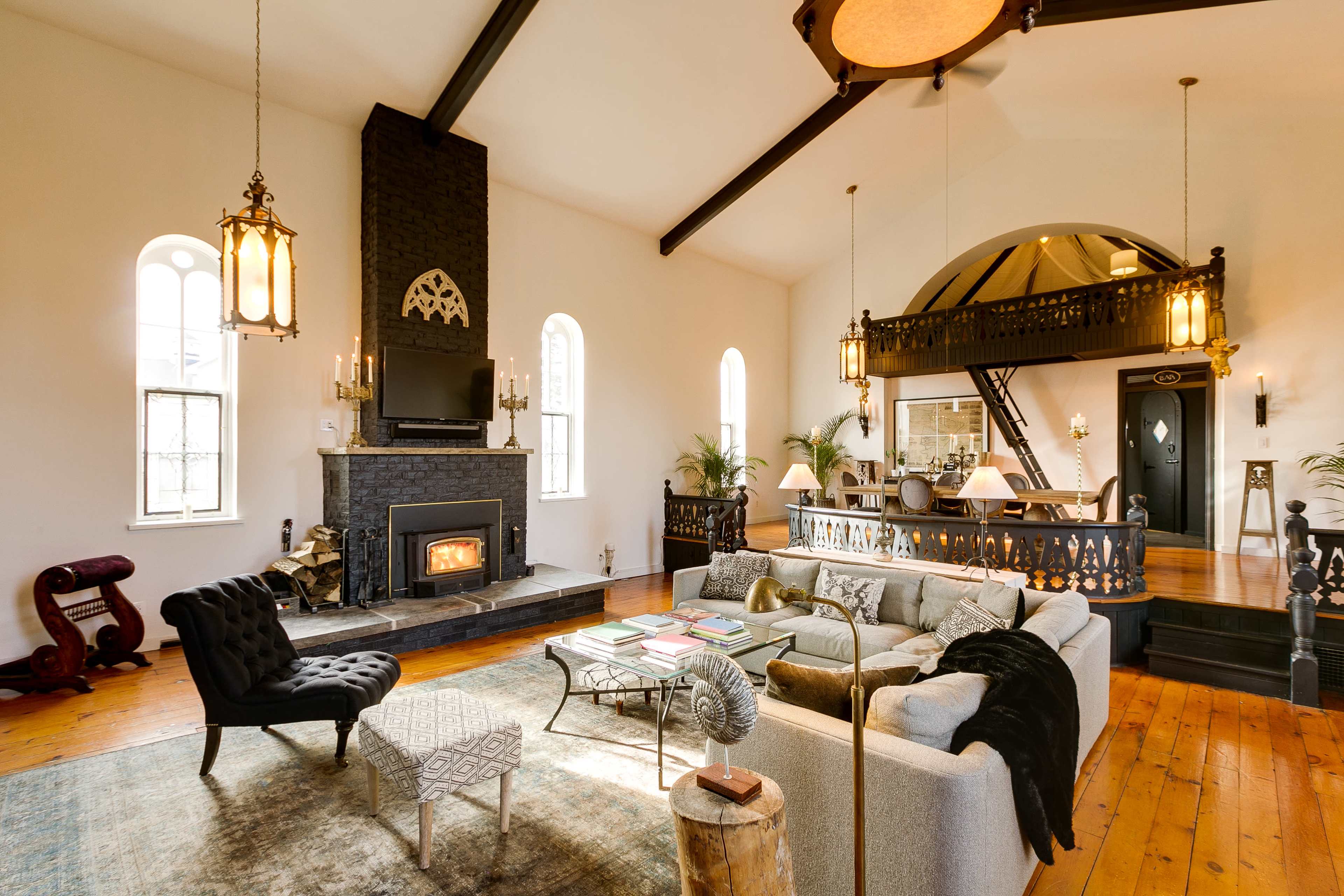 The image shows a spacious living room with a central fireplace, high ceilings, and a mix of comfortable seating arrangements.