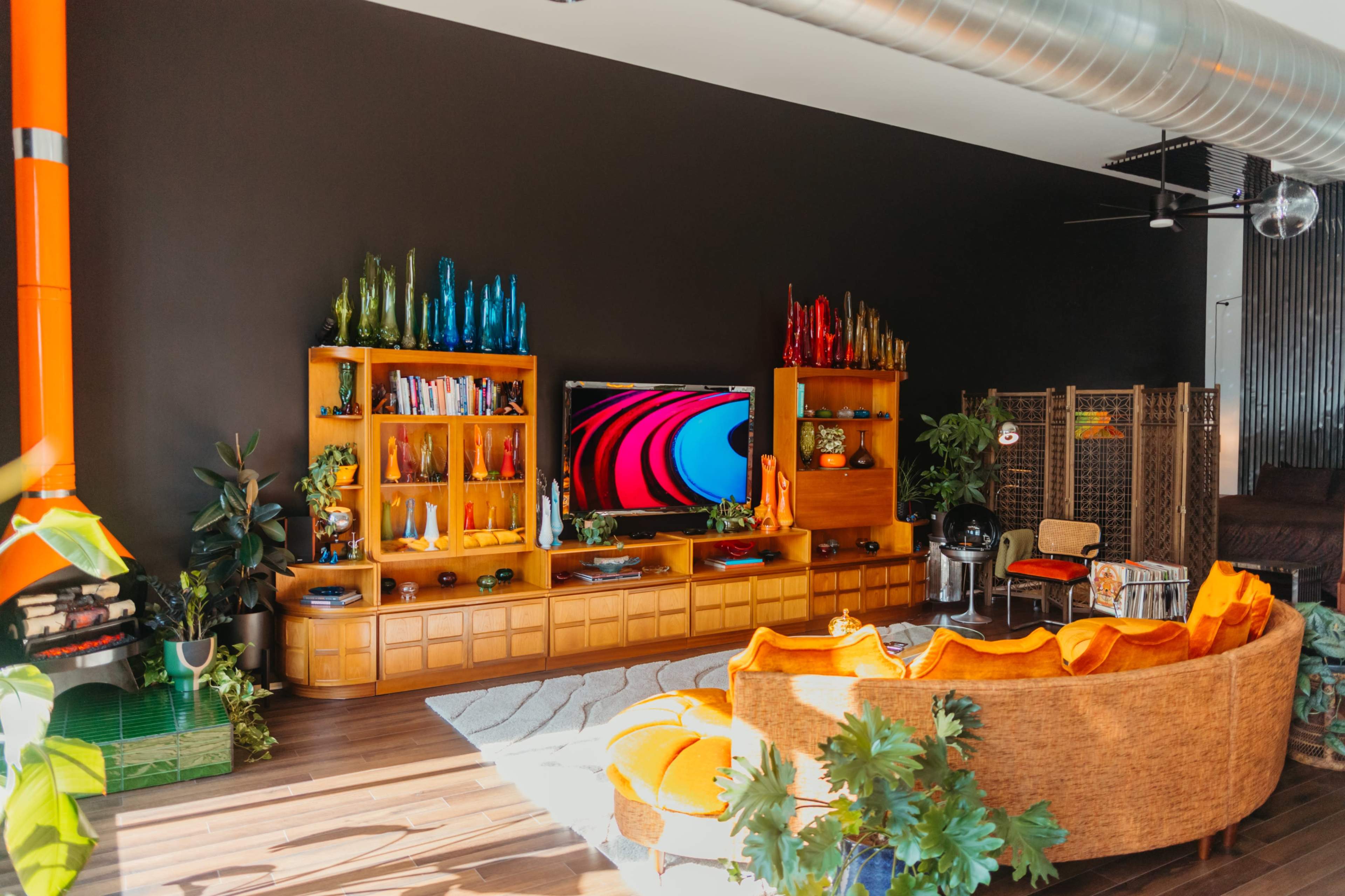 A modern living room features a large wall-mounted TV flanked by wooden shelves adorned with colorful decorative bottles, surrounded by a curved orange sofa and green plants.