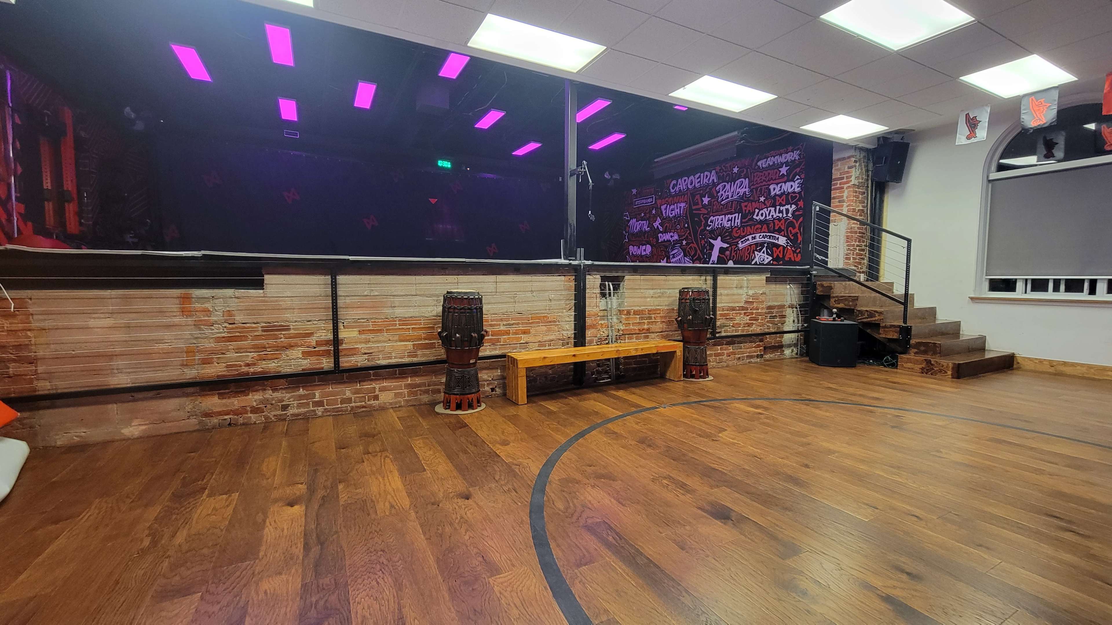 The image shows a spacious room with wooden flooring, a stage area with colorful lighting, and a backdrop featuring graffiti-style art, along with two drum-like instruments and a bench.