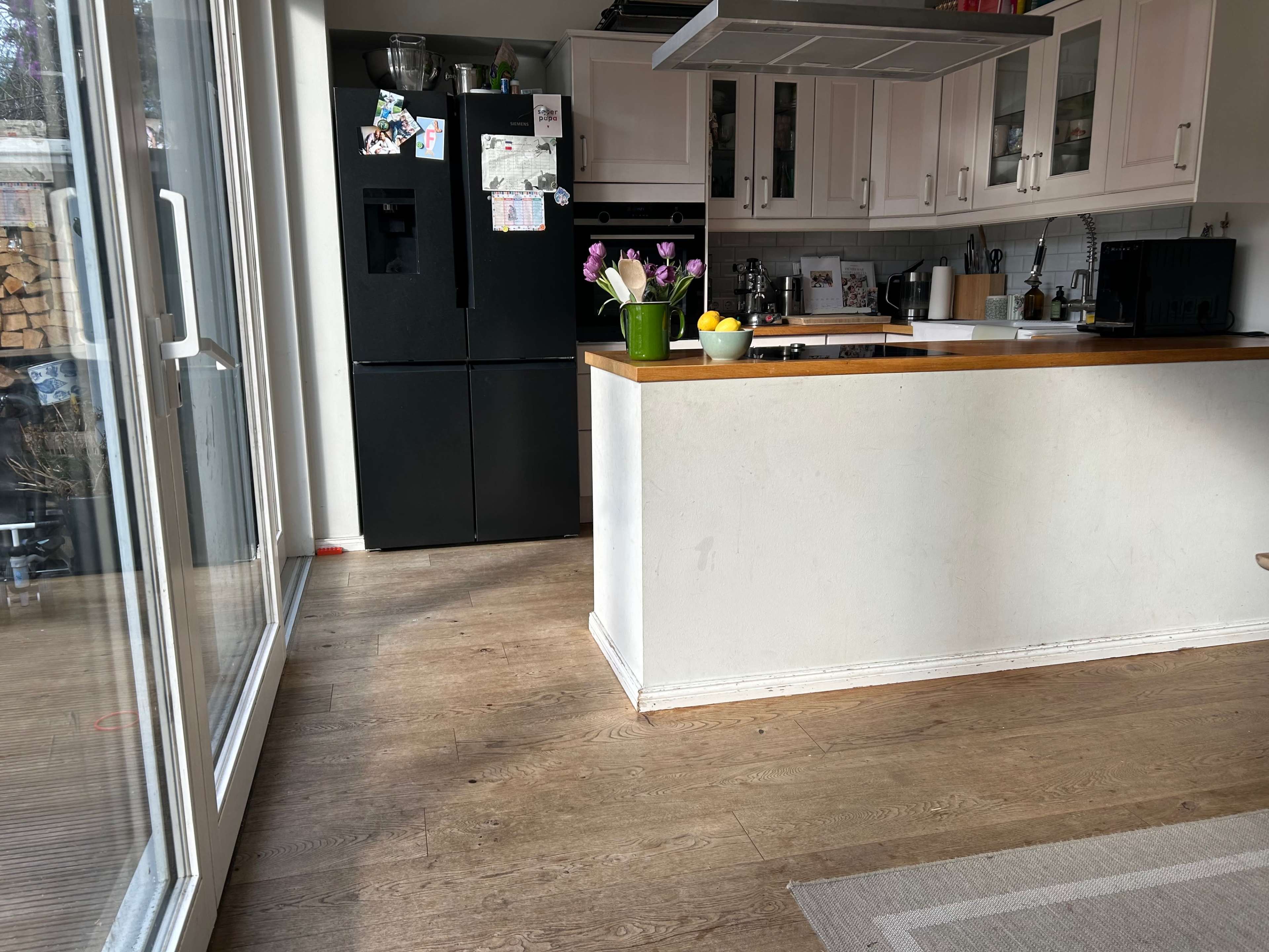 The image shows a kitchen with light-colored cabinets, a black refrigerator, a bowl of fruit, and a vase of flowers on a countertop.