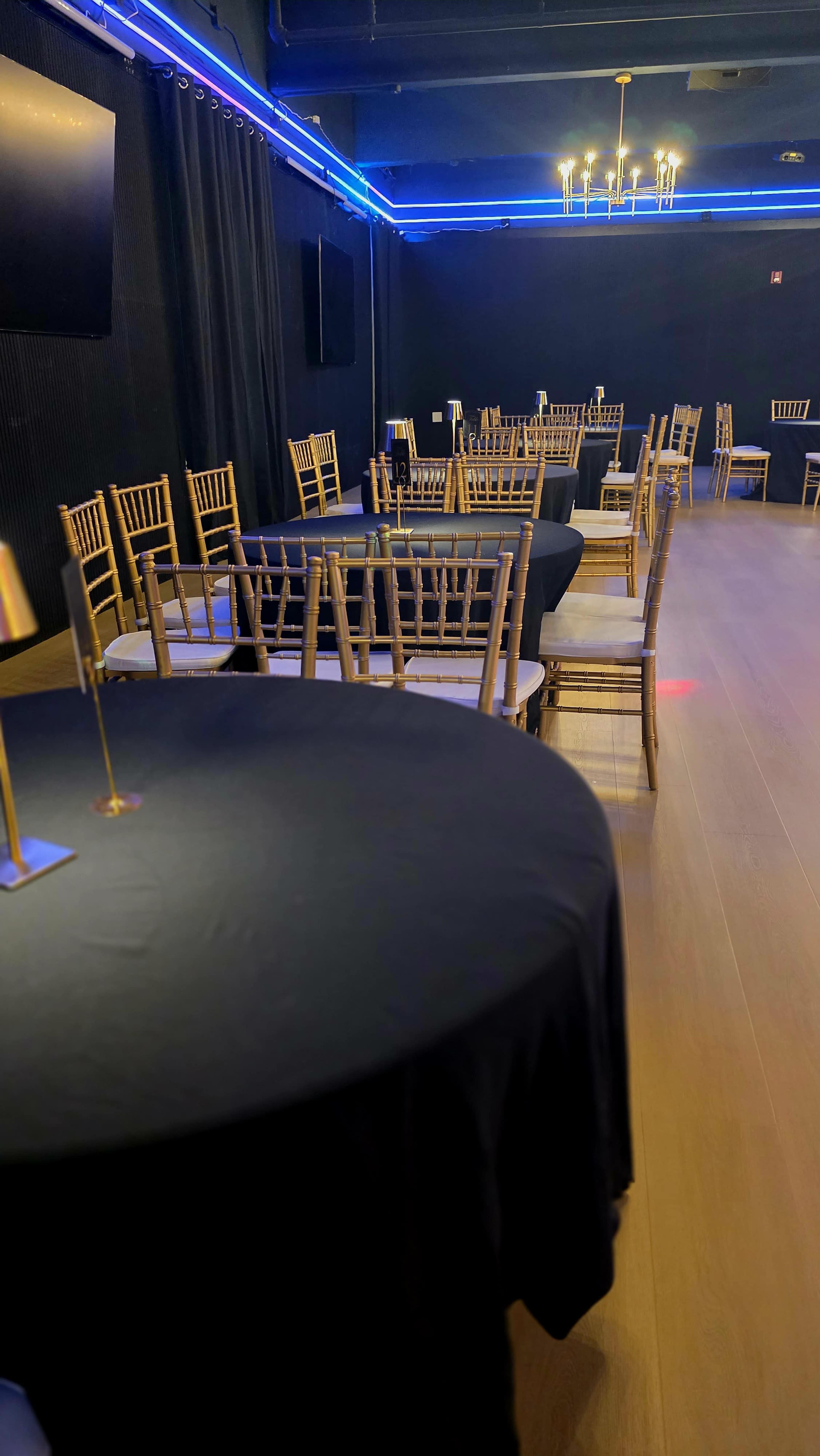 The image shows a spacious event room with rectangular tables covered in black cloth and gold chiavari chairs arranged around them, illuminated by blue lighting along the walls.