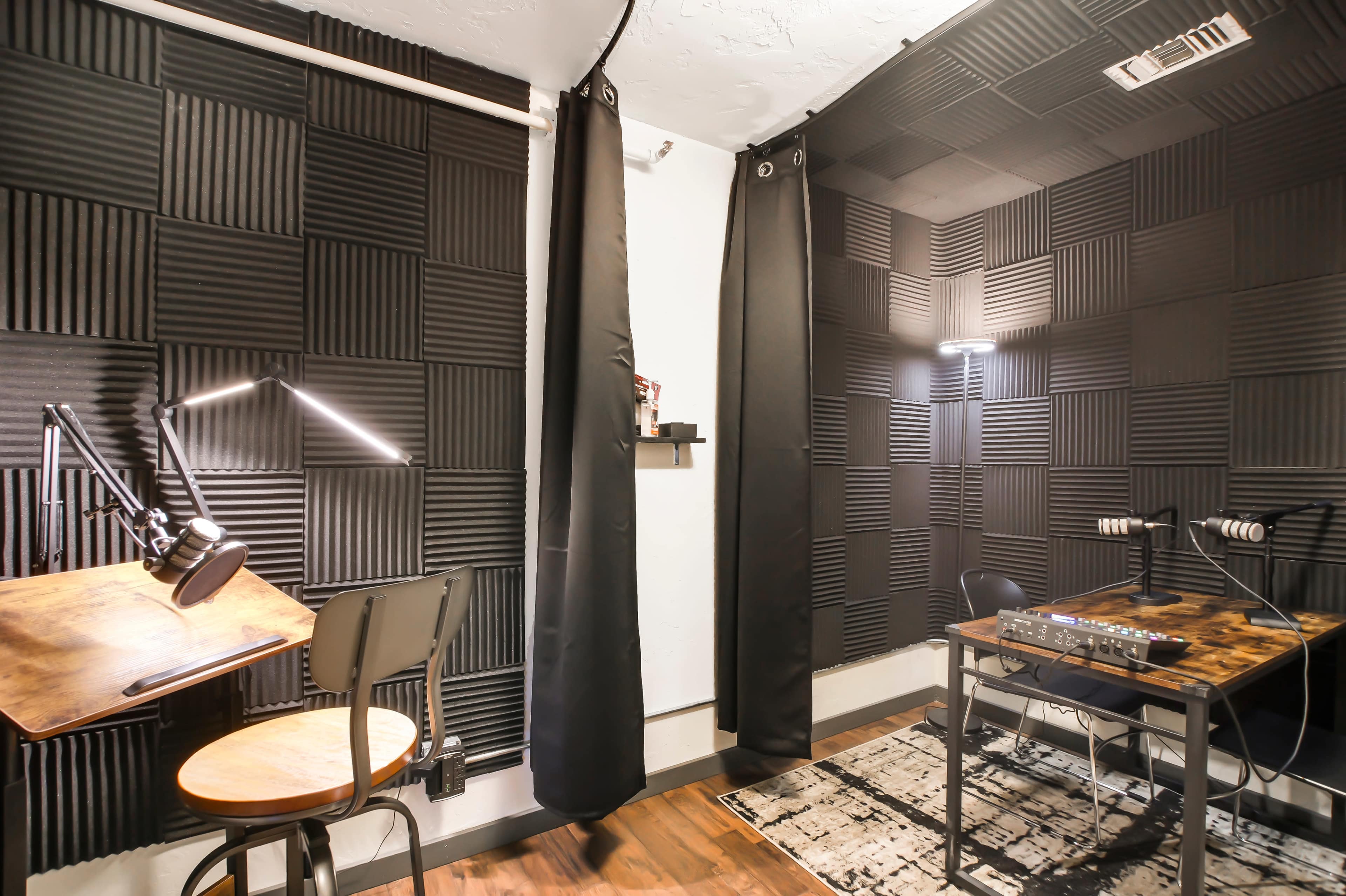 A soundproof recording studio featuring black acoustic foam panels on the walls, two desks with microphones, and a rug on the floor.