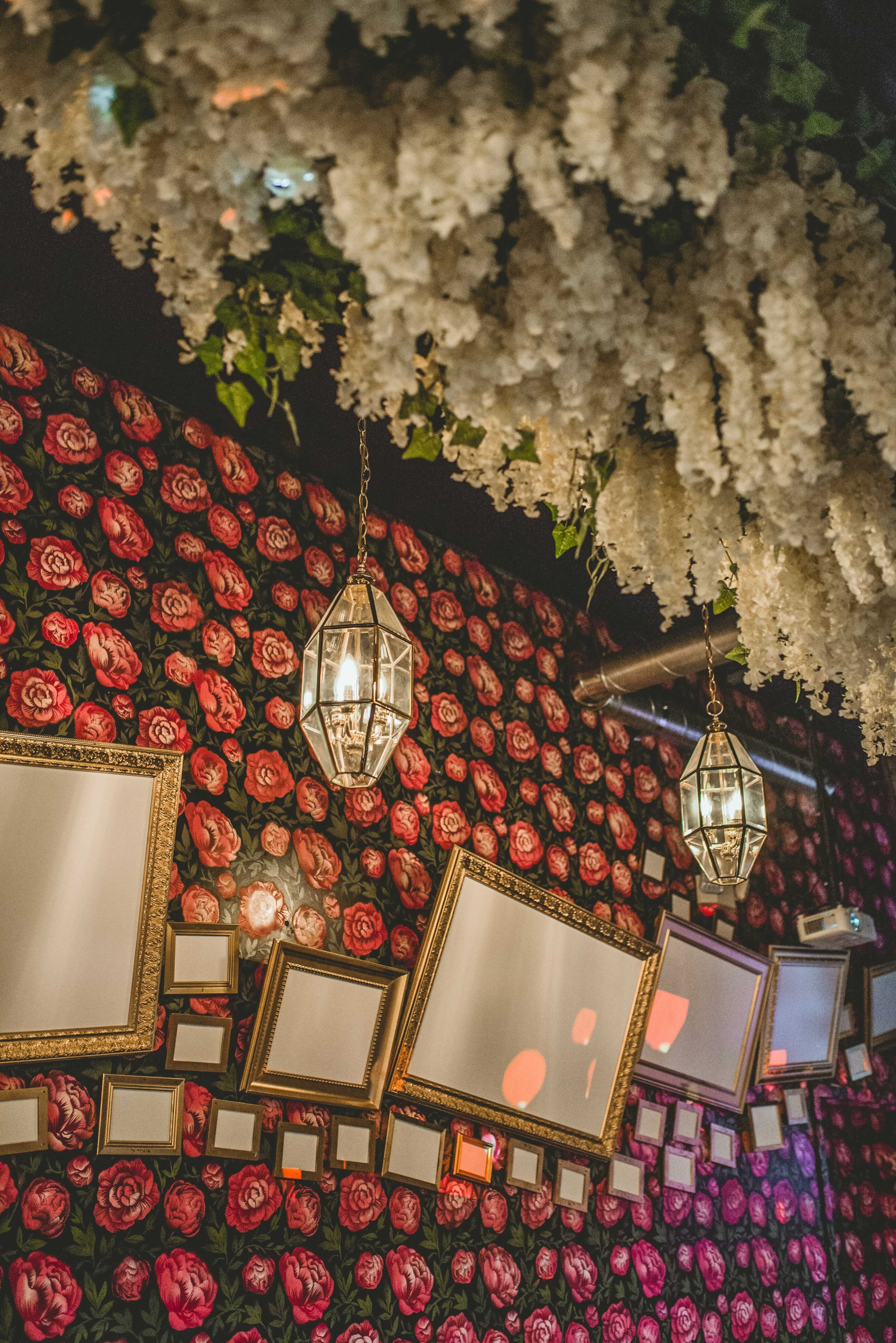 The scene features a wall adorned with floral wallpaper, multiple ornate mirrors, and hanging light fixtures beneath a cascade of artificial flowers.