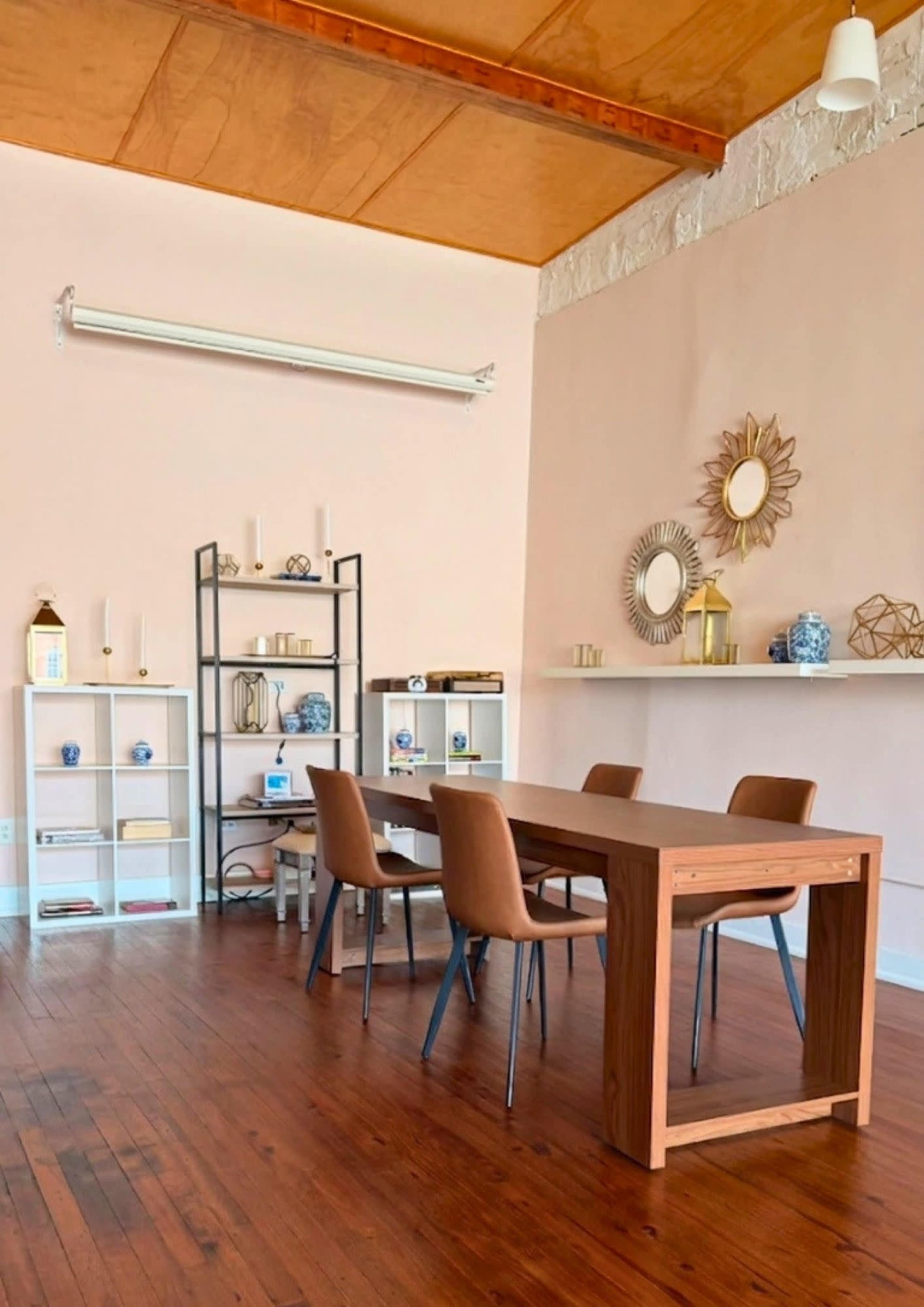 The image shows a bright room featuring a wooden table surrounded by four chairs, along with shelves displaying decorative items against pale pink walls.