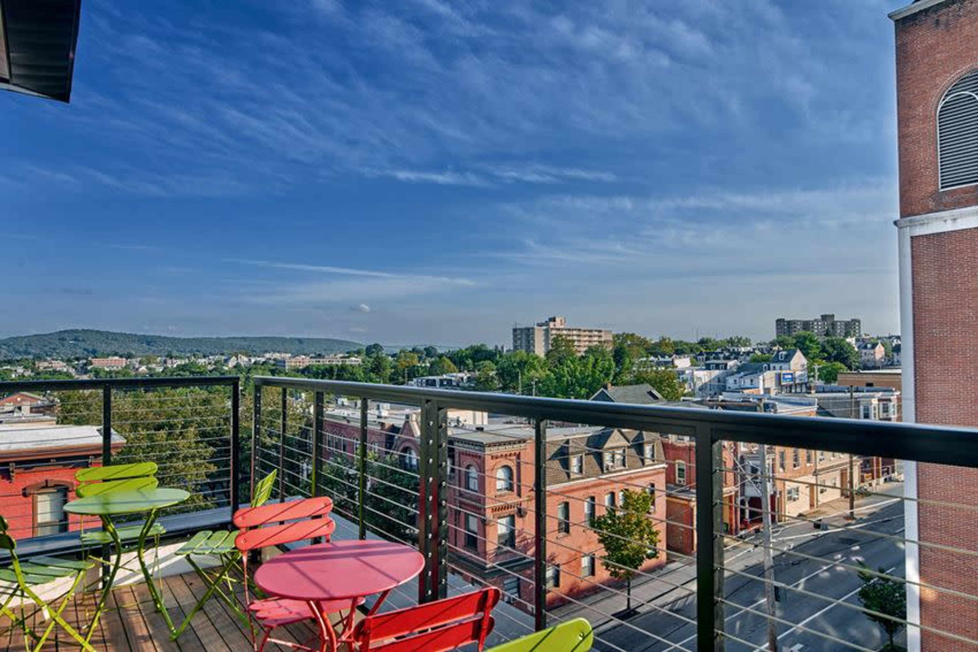 A rooftop balcony features colorful seating and offers a view of a cityscape with buildings and a distant mountain under a clear sky.