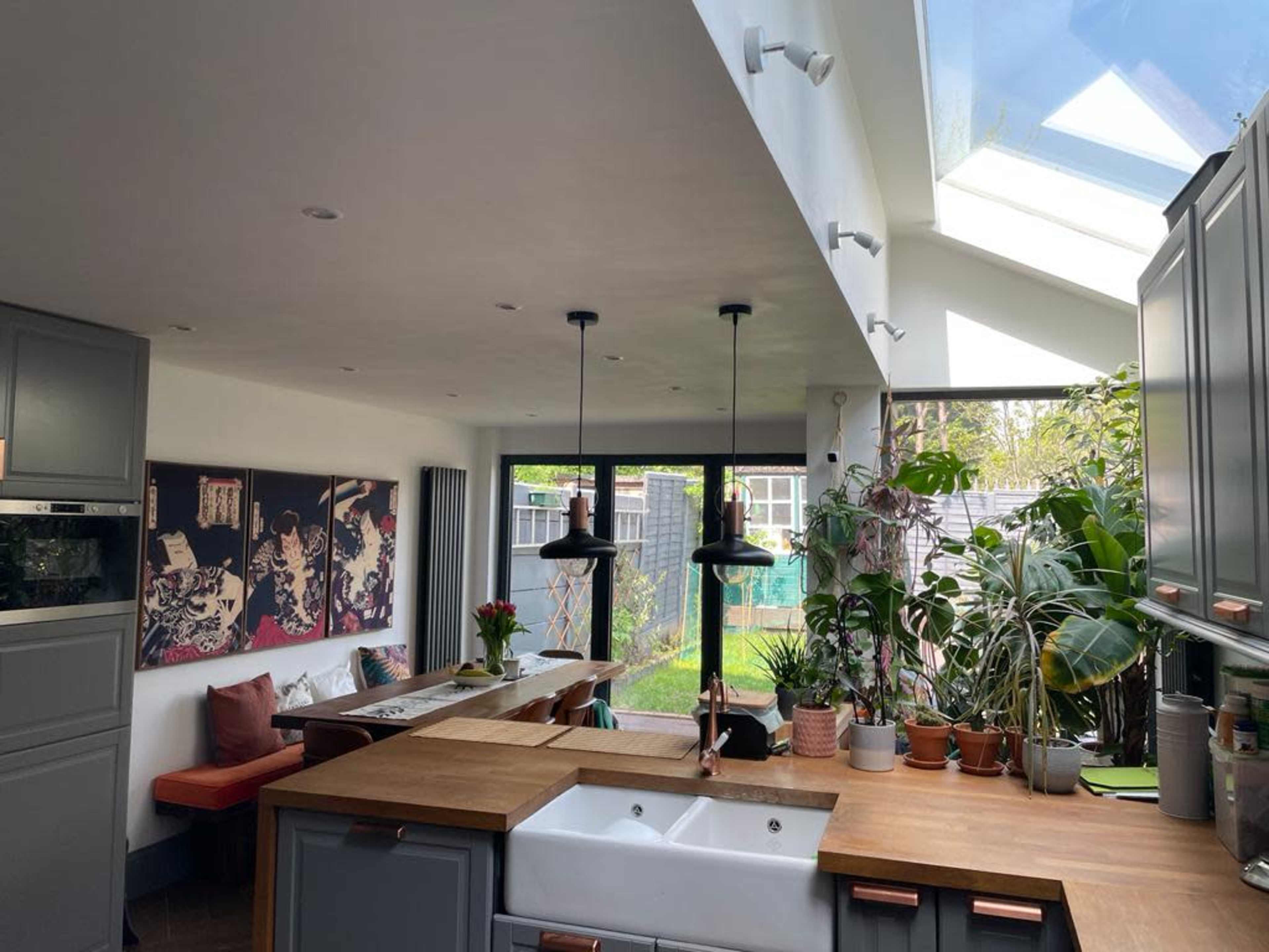 A modern kitchen and dining area featuring a central island with a farmhouse sink, surrounded by plants and large windows that provide natural light.