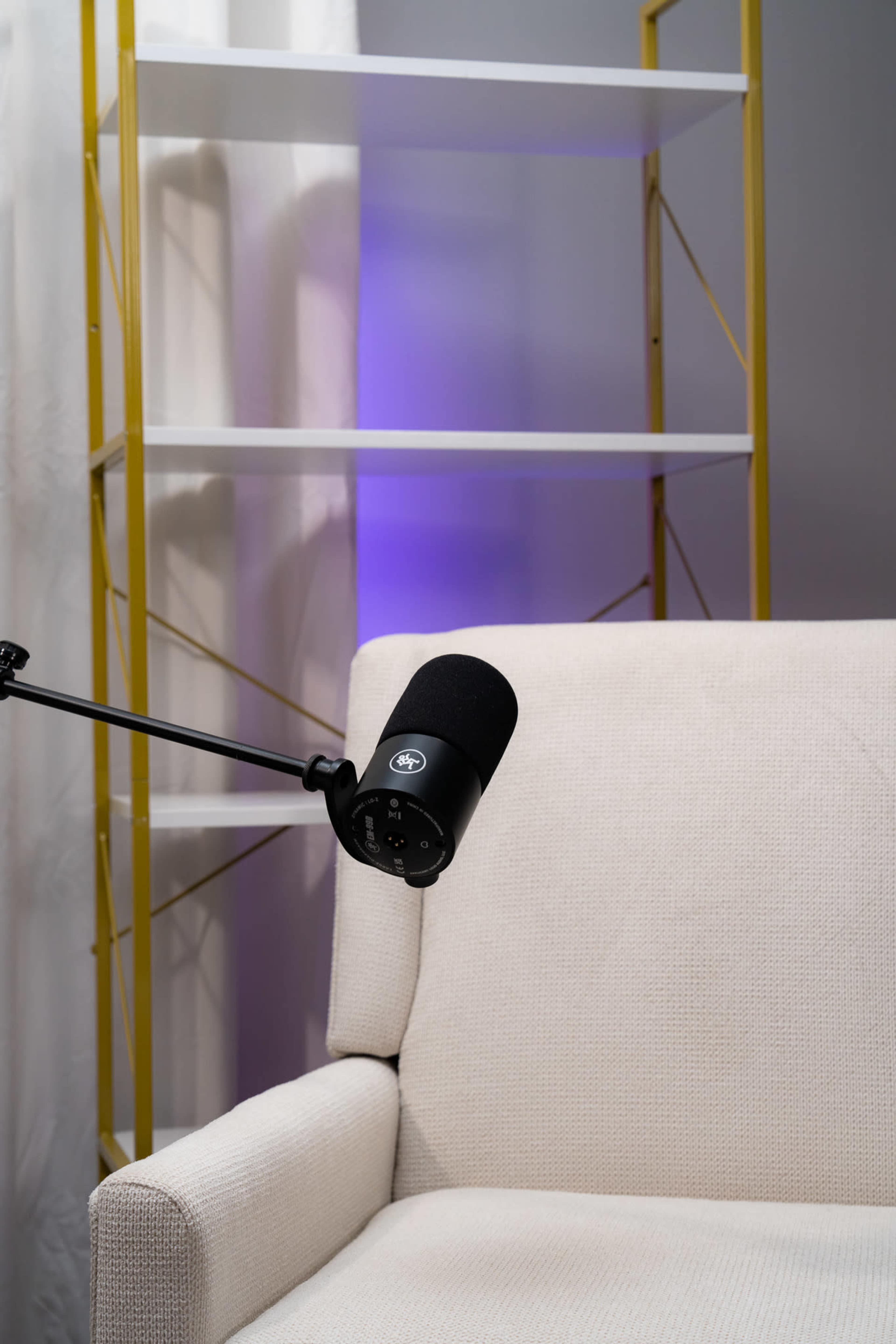 A microphone is mounted on an adjustable arm next to a white couch in a well-lit room with a yellow shelving unit.