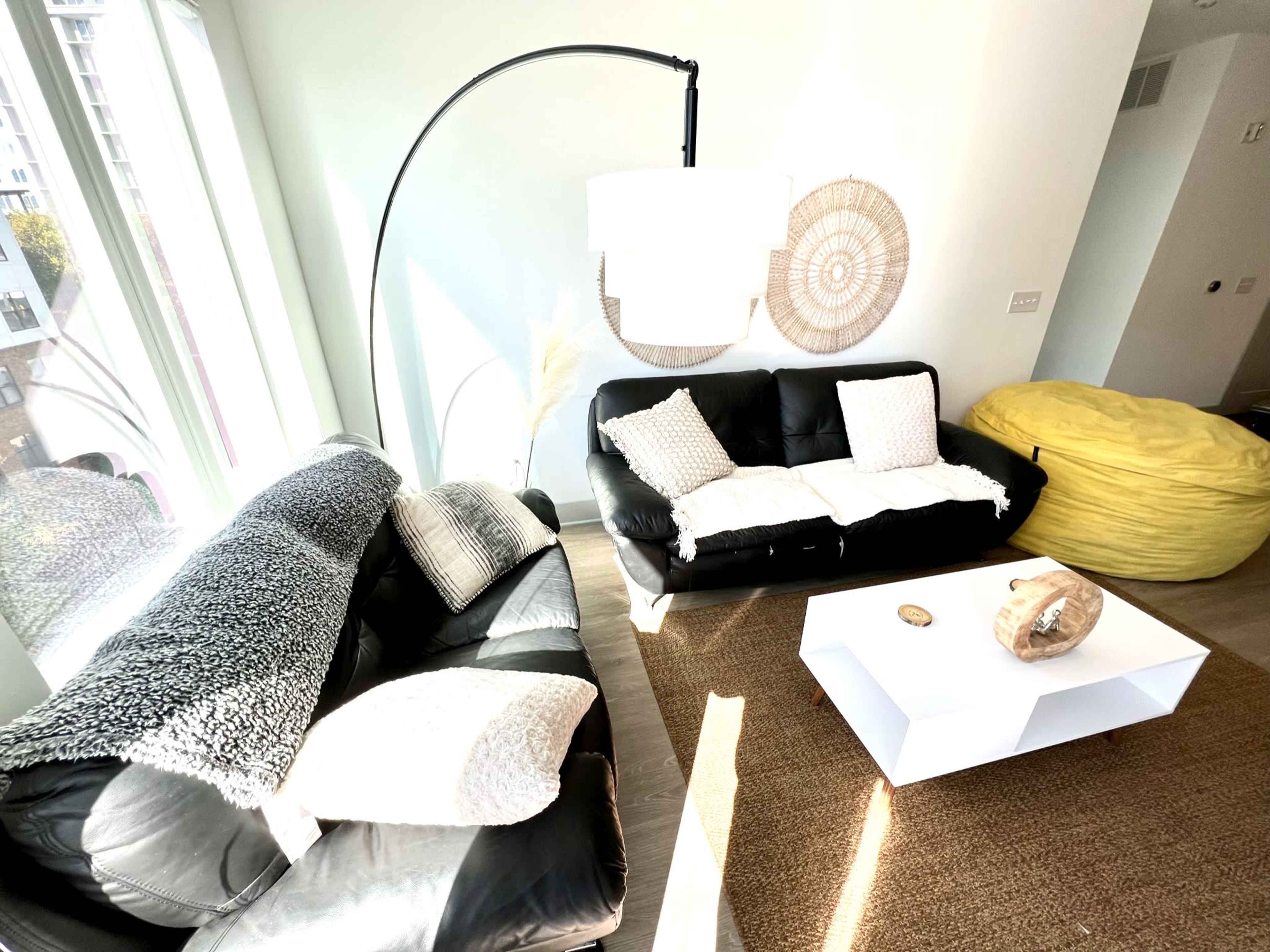 A bright, modern living room features two black sofas, a yellow bean bag chair, a white coffee table, and decorative wall elements.