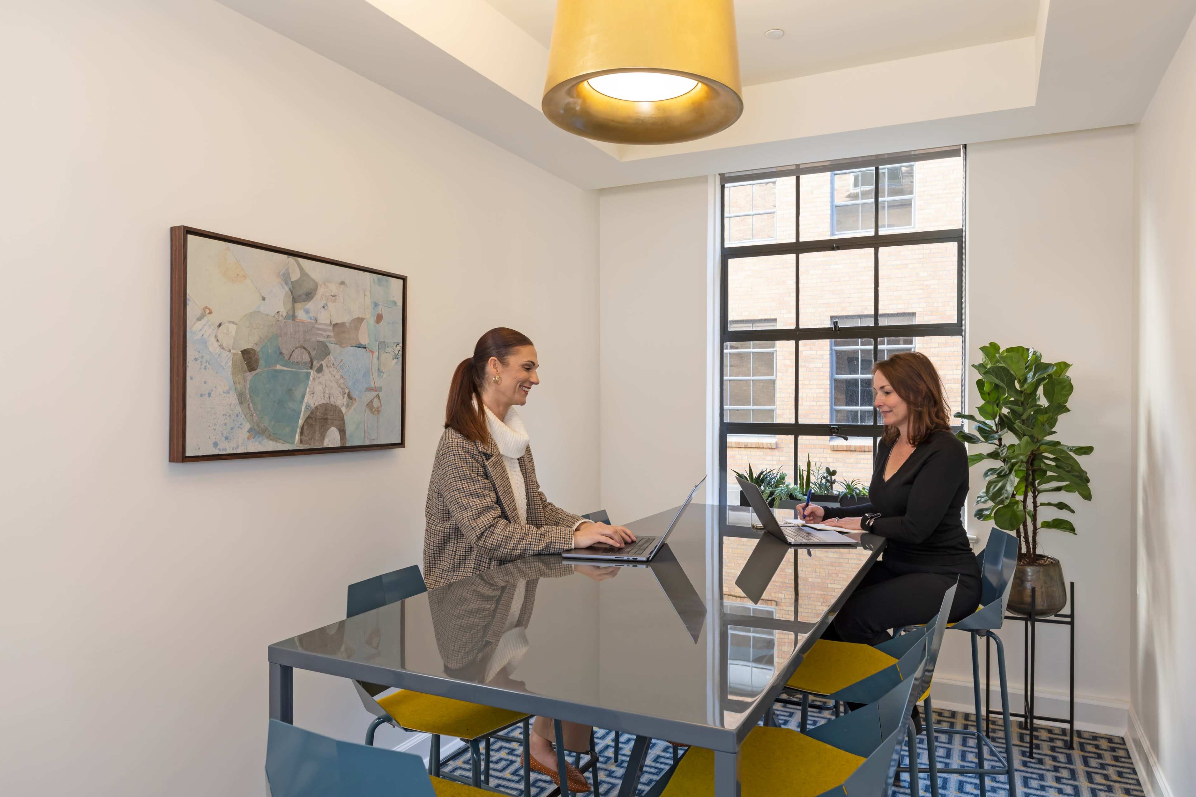 Two women are seated at a modern table in a well-lit office space, engaging in a discussion while using laptops.