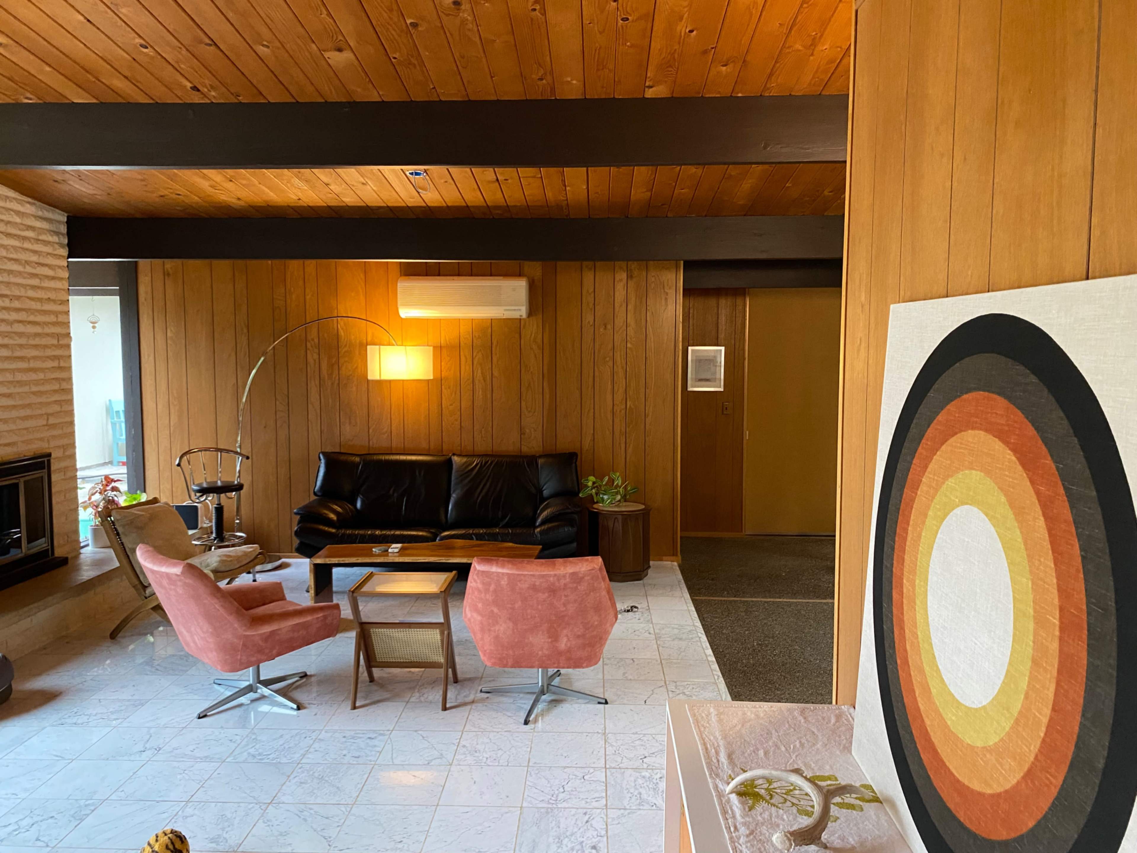A mid-century modern living room featuring a black leather sofa, two pink chairs, and a round coffee table on a marble floor.