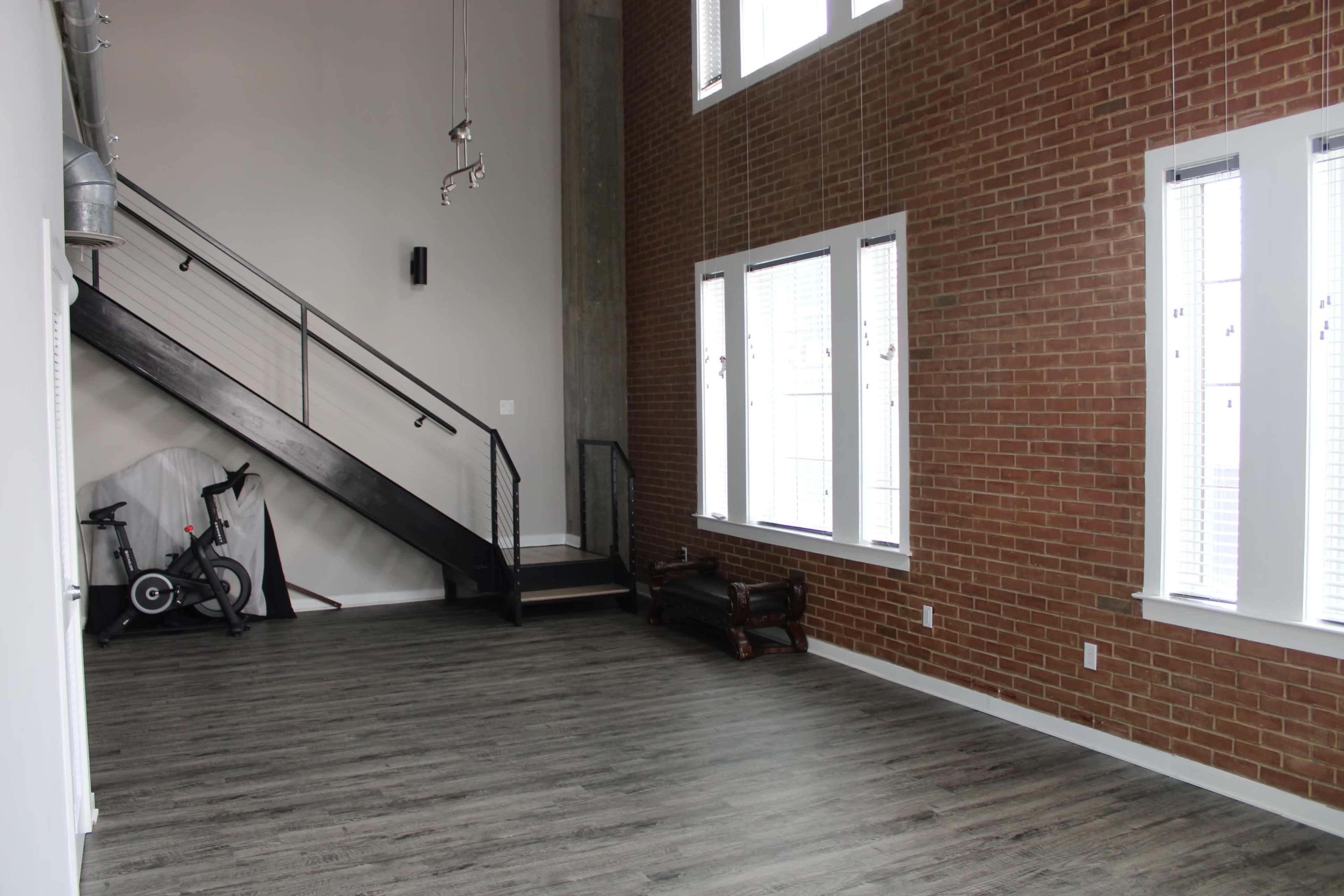 A spacious room with a high ceiling, featuring a brick wall, large windows, a staircase, and a bicycle in the corner.