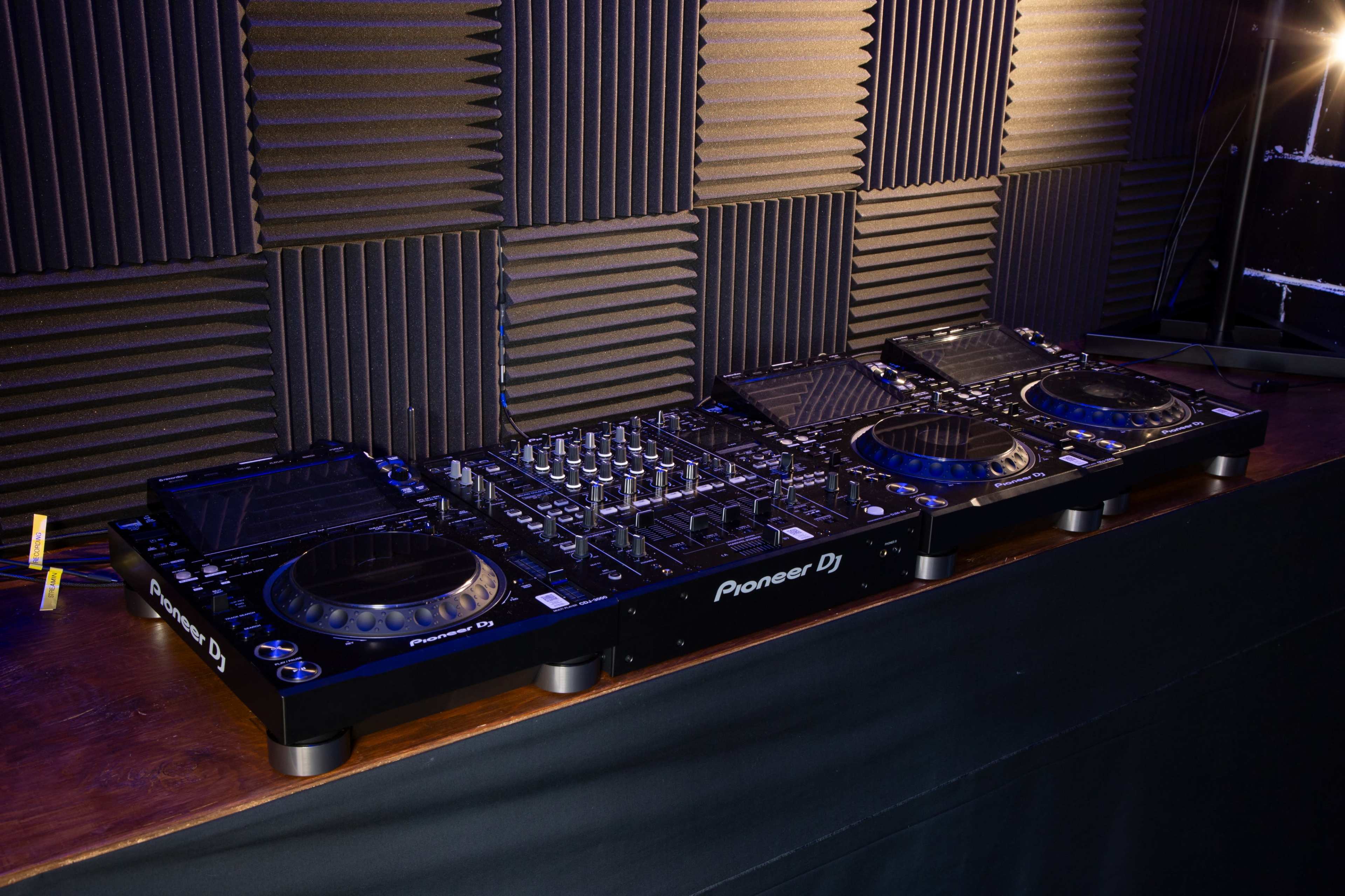 A Pioneer DJ setup is placed on a wooden table in a room with soundproof foam panels on the walls.