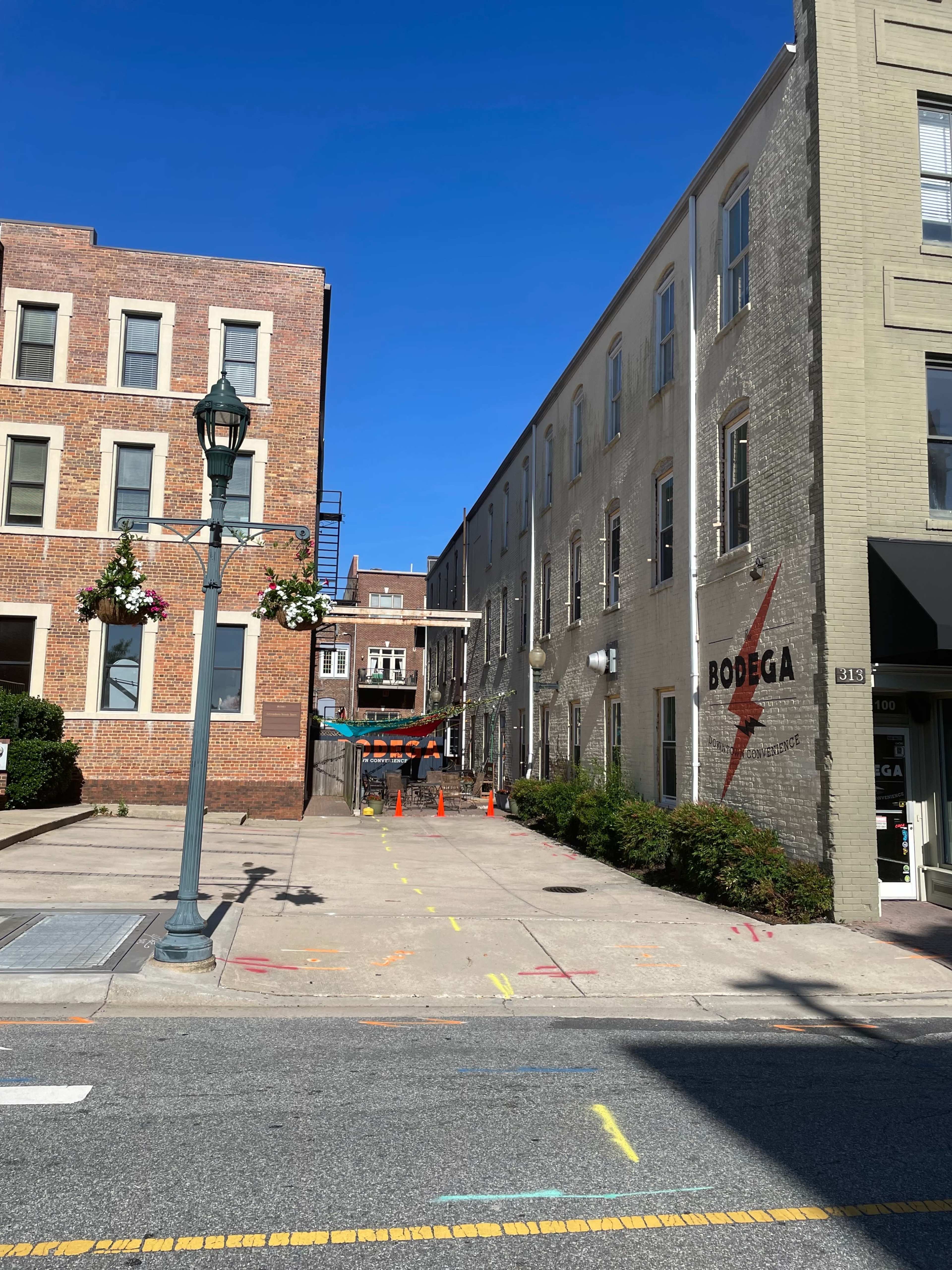 A narrow alleyway between brick buildings features a sign for a bodega and is marked by construction cones and colorful chalk lines on the pavement.
