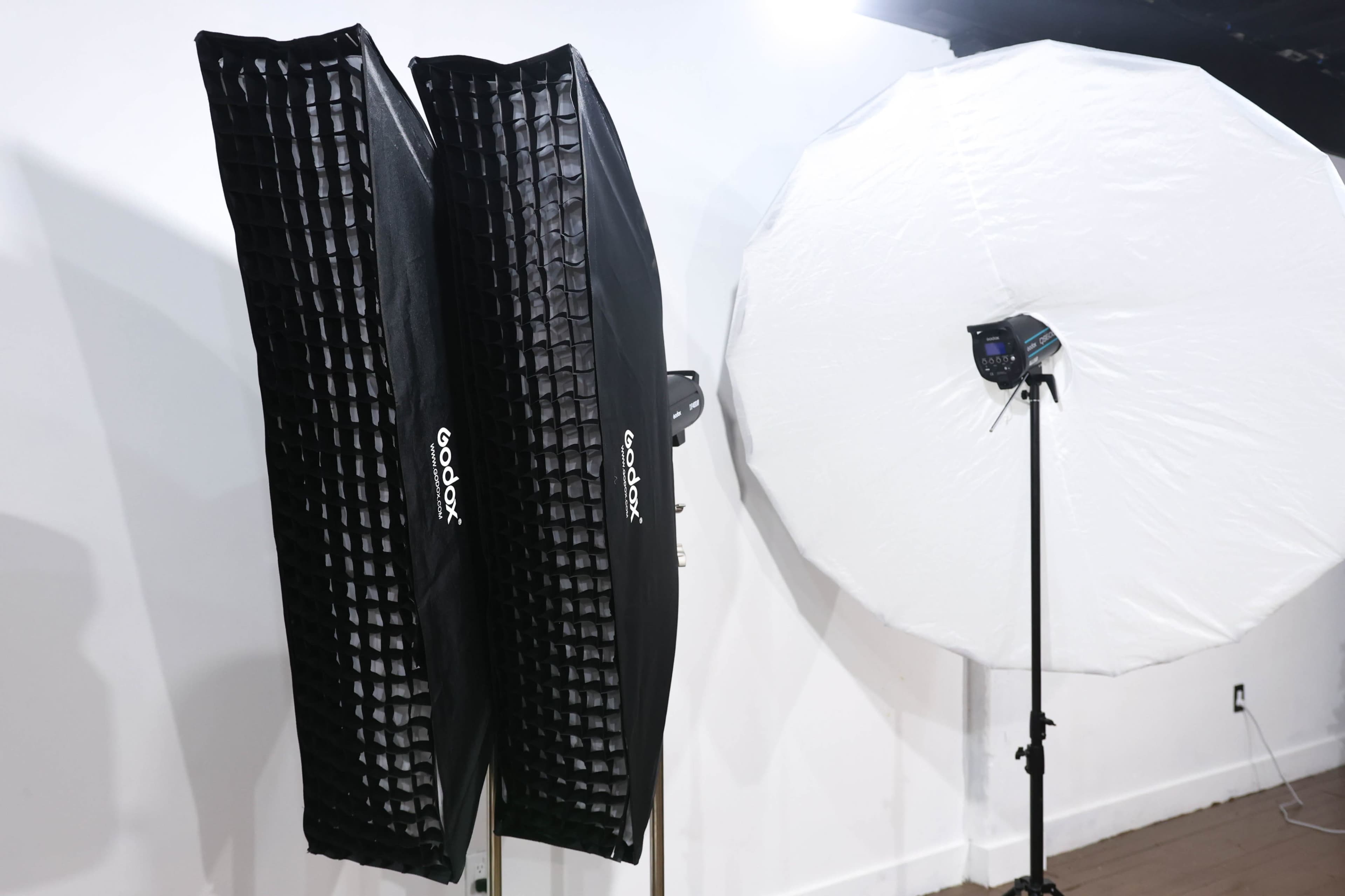 Two black softbox lighting modifiers alongside a large white umbrella, all set up in a photography studio.
