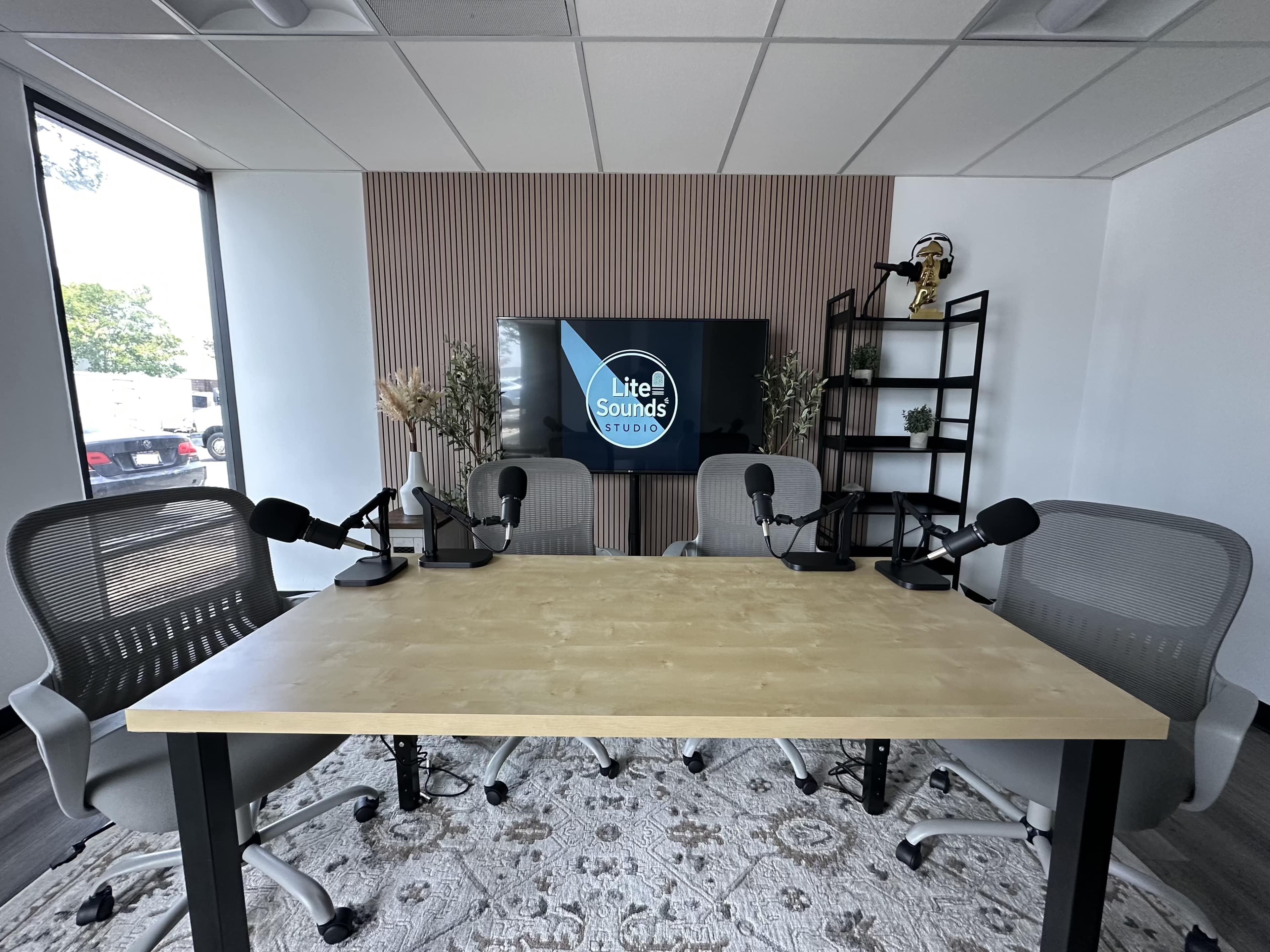 A modern podcast recording studio features a table with microphones, four chairs, decorative plants, and a wall-mounted screen displaying the studio's name.