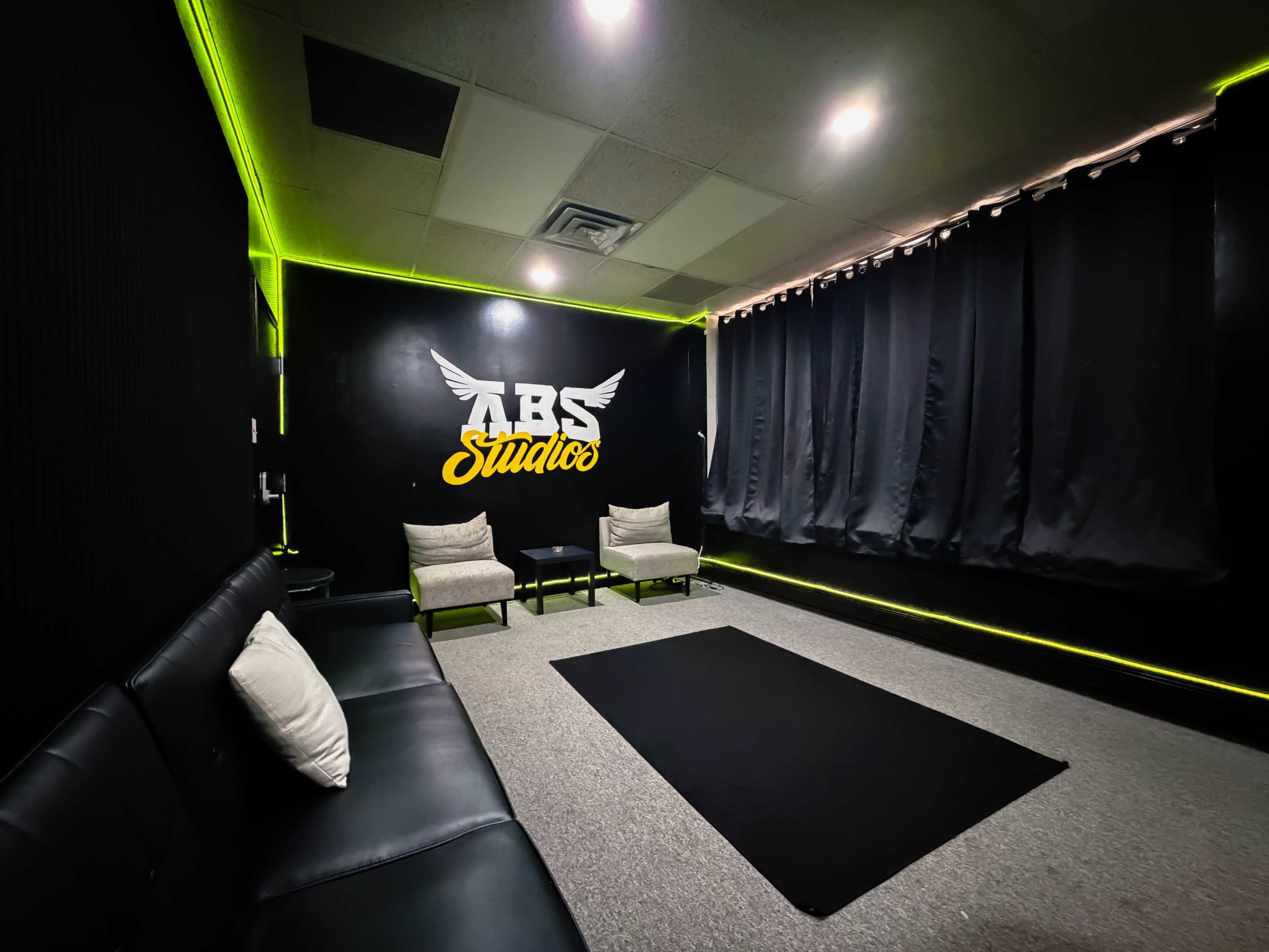 The image shows a modern, black-walled studio space with two chairs, a couch, and a black rug, illuminated by green LED lights along the walls.
