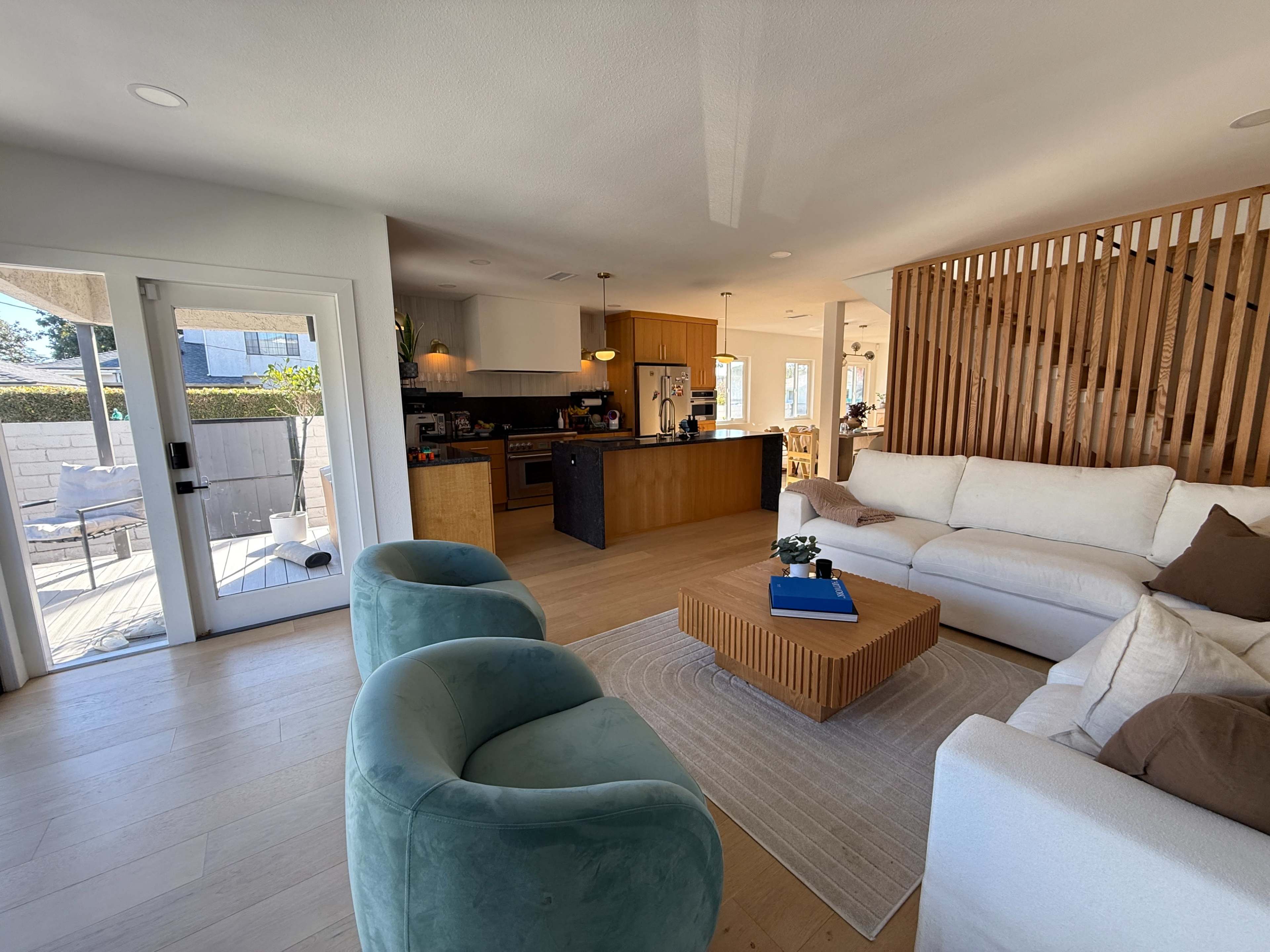 The image shows a modern living room with a light color palette, featuring two teal chairs, a wooden coffee table, and an open layout leading to a kitchen area.