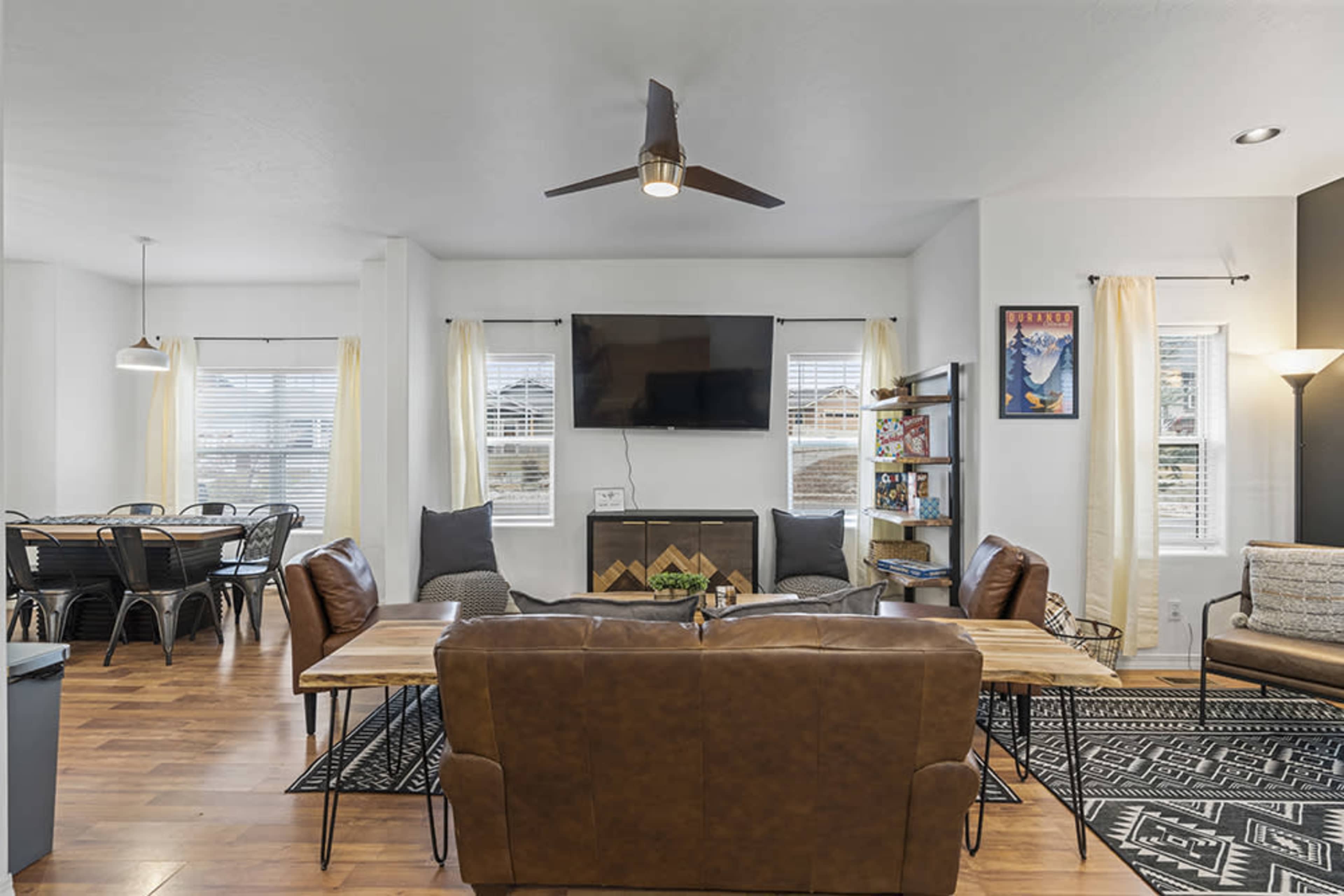 A modern living area with a flat-screen TV mounted above a fireplace, surrounded by brown leather couches, a wooden coffee table, and a dining area with black metal chairs in the background.