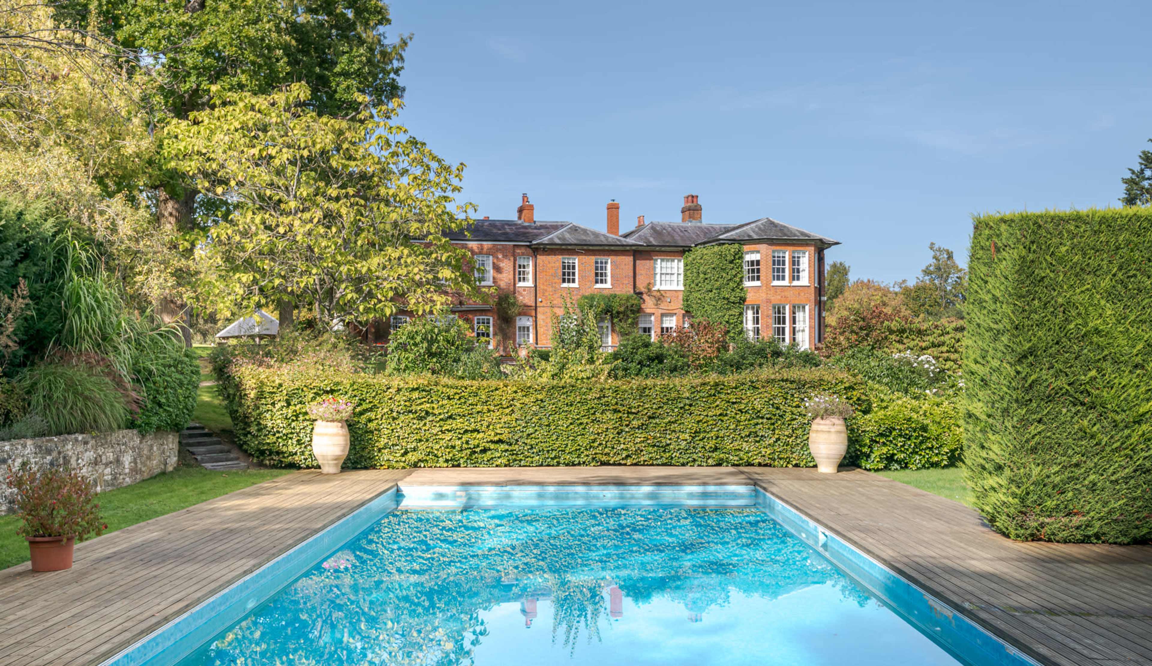 A clear swimming pool with a wooden deck is situated in front of a large red-brick mansion surrounded by lush greenery.