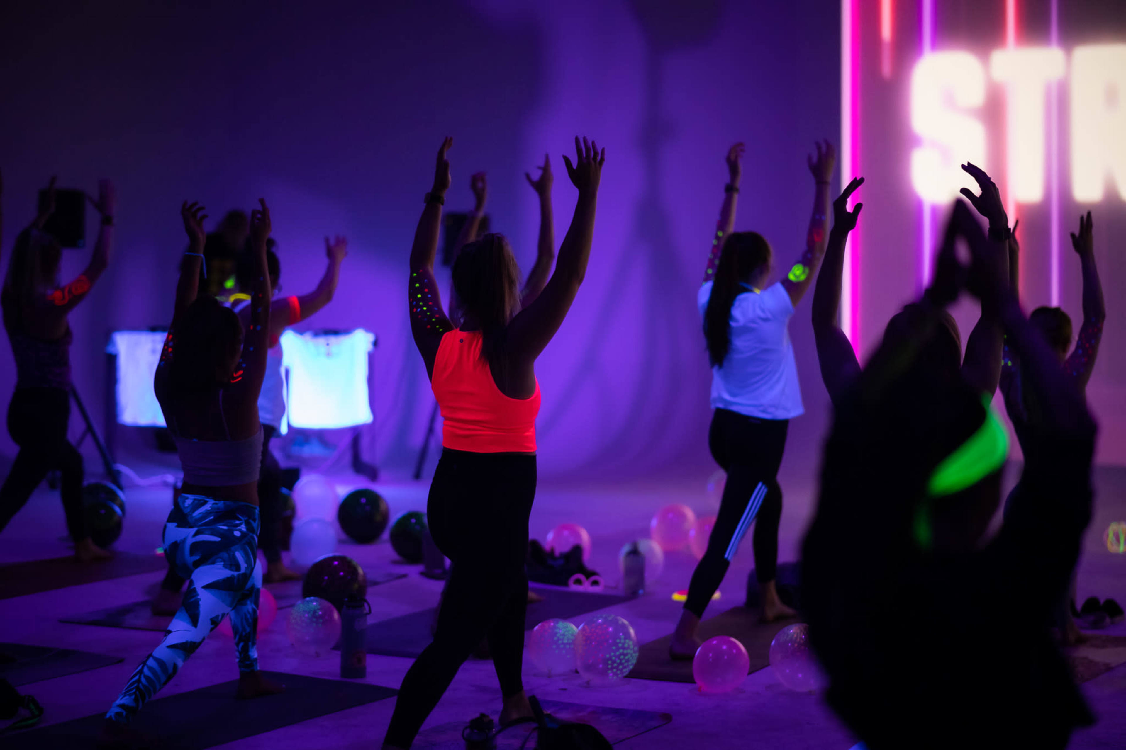 A group of individuals is practicing yoga in a dimly lit studio with vibrant neon lights and colorful exercise mats.