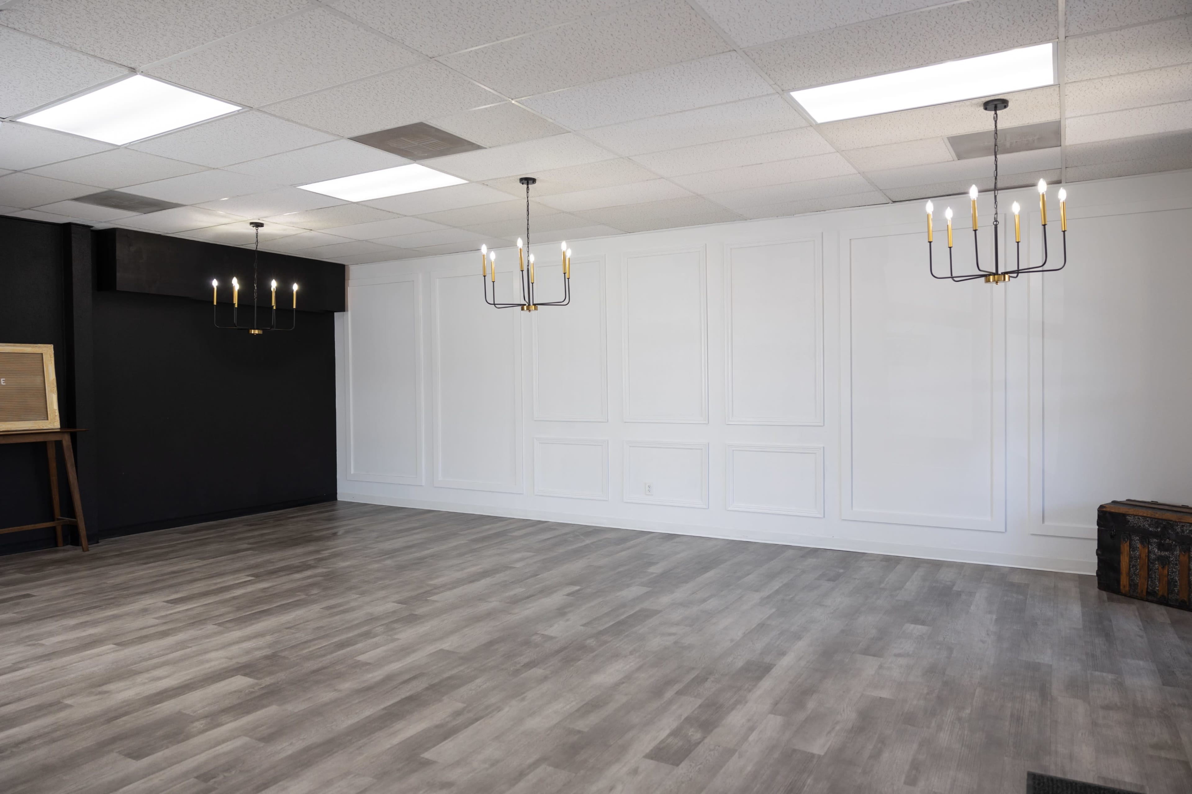 A spacious, empty room with a light-colored floor, white walls with decorative paneling, and three chandeliers hanging from the ceiling.