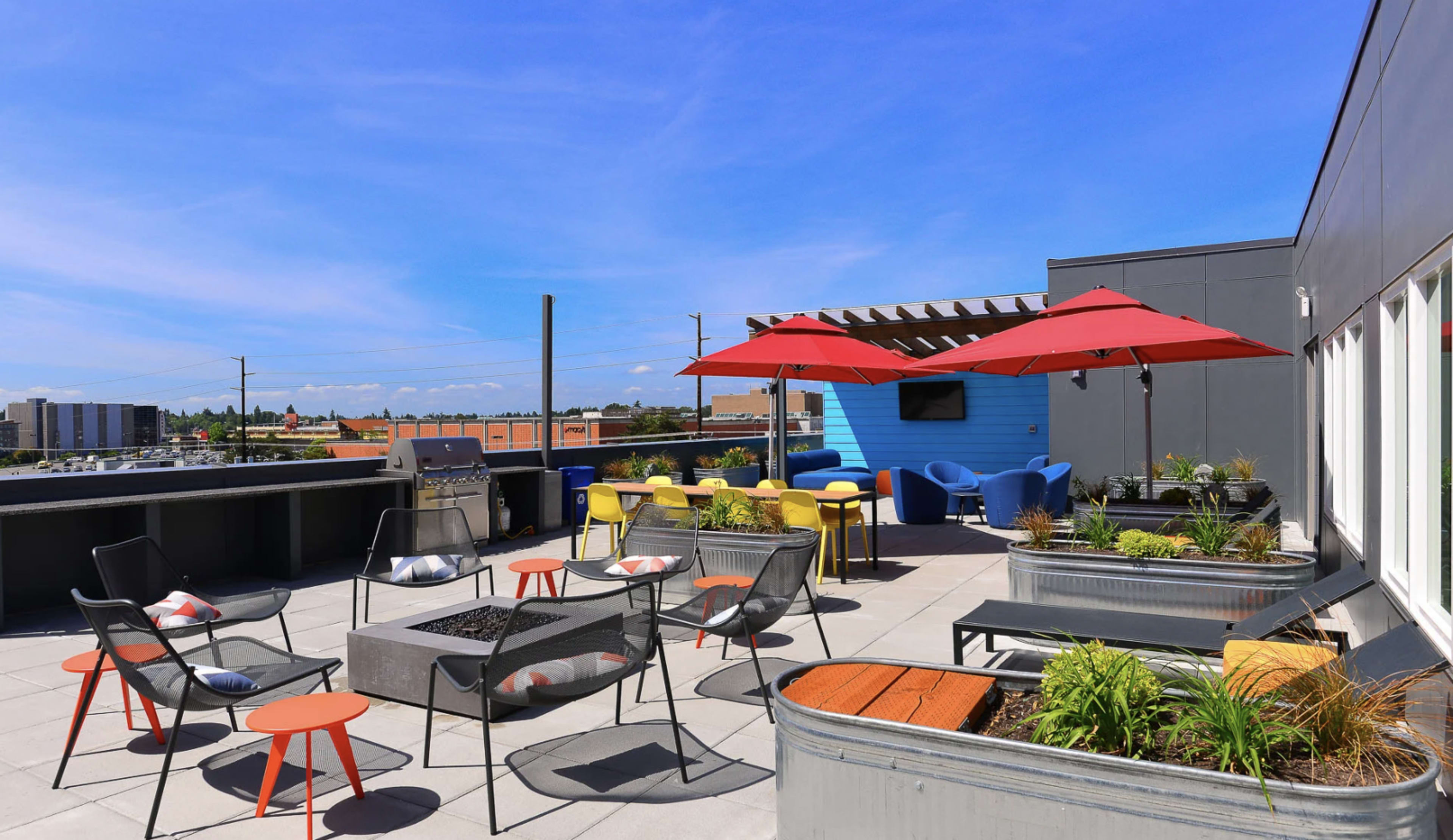 The image shows a rooftop terrace featuring colorful seating, a barbecue area, and planters, with a clear blue sky in the background.