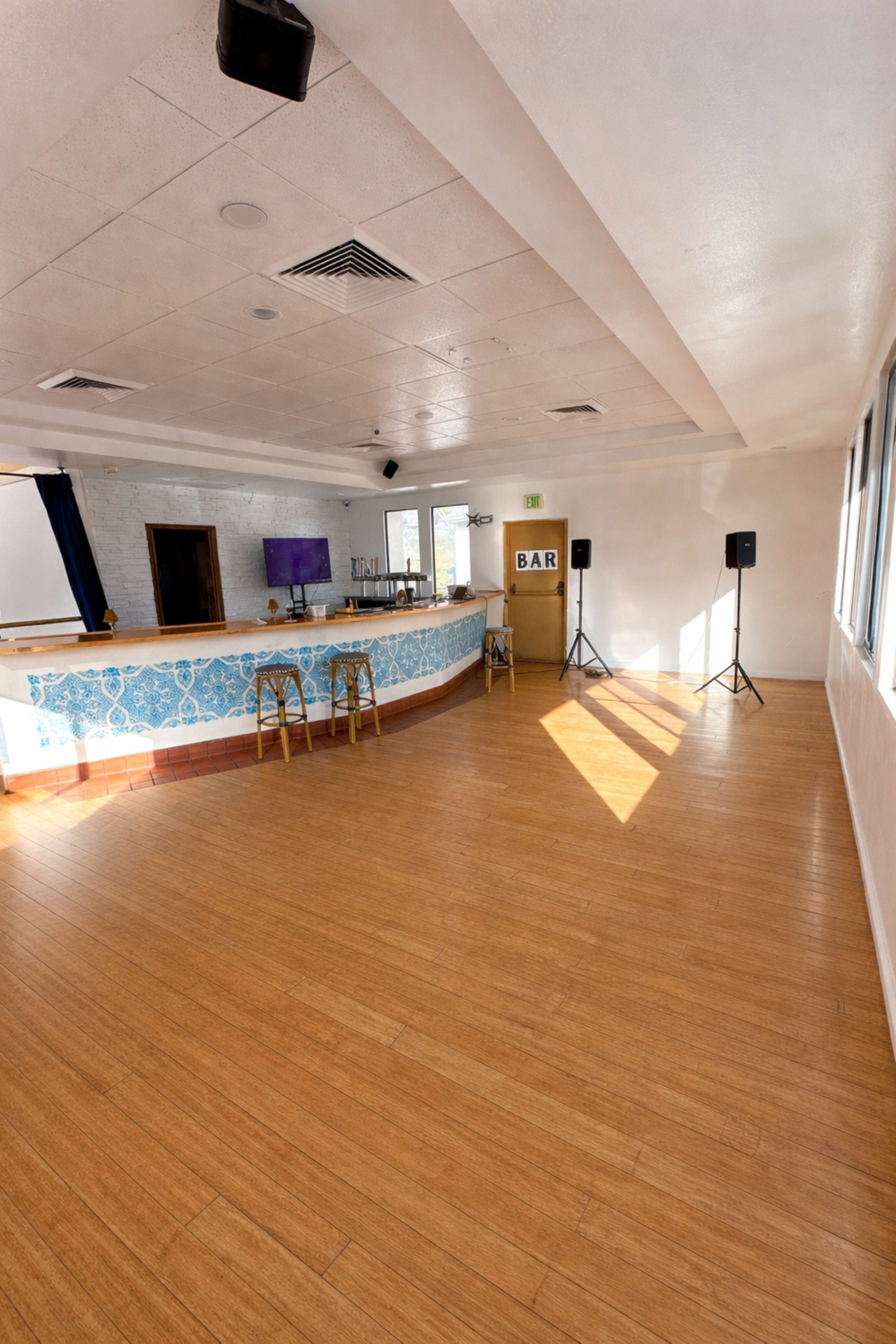 The image shows an empty indoor space featuring a bar with a blue patterned front, wooden flooring, and speakers set up in the corners.