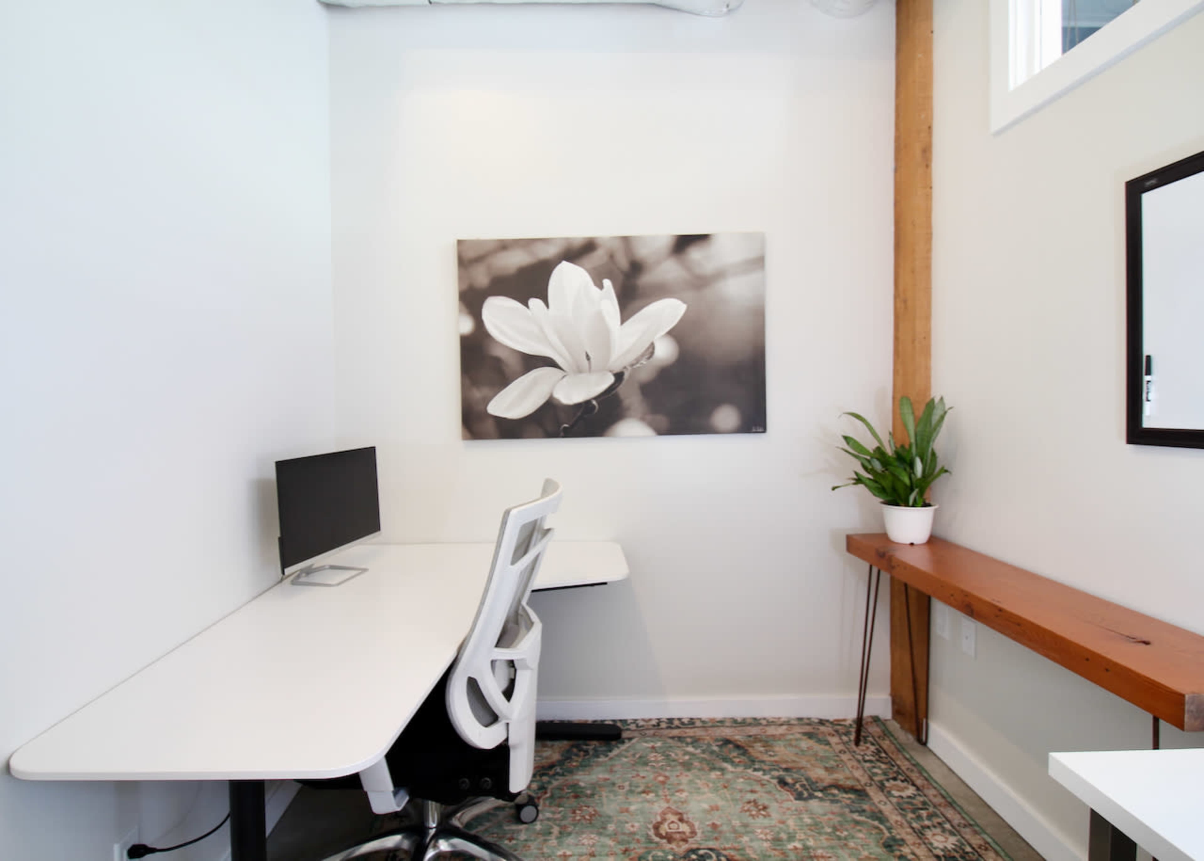 The image shows a clean, minimalistic office space featuring a desk with a computer monitor, a wooden shelf against the wall, a potted plant, and a black-and-white floral artwork.