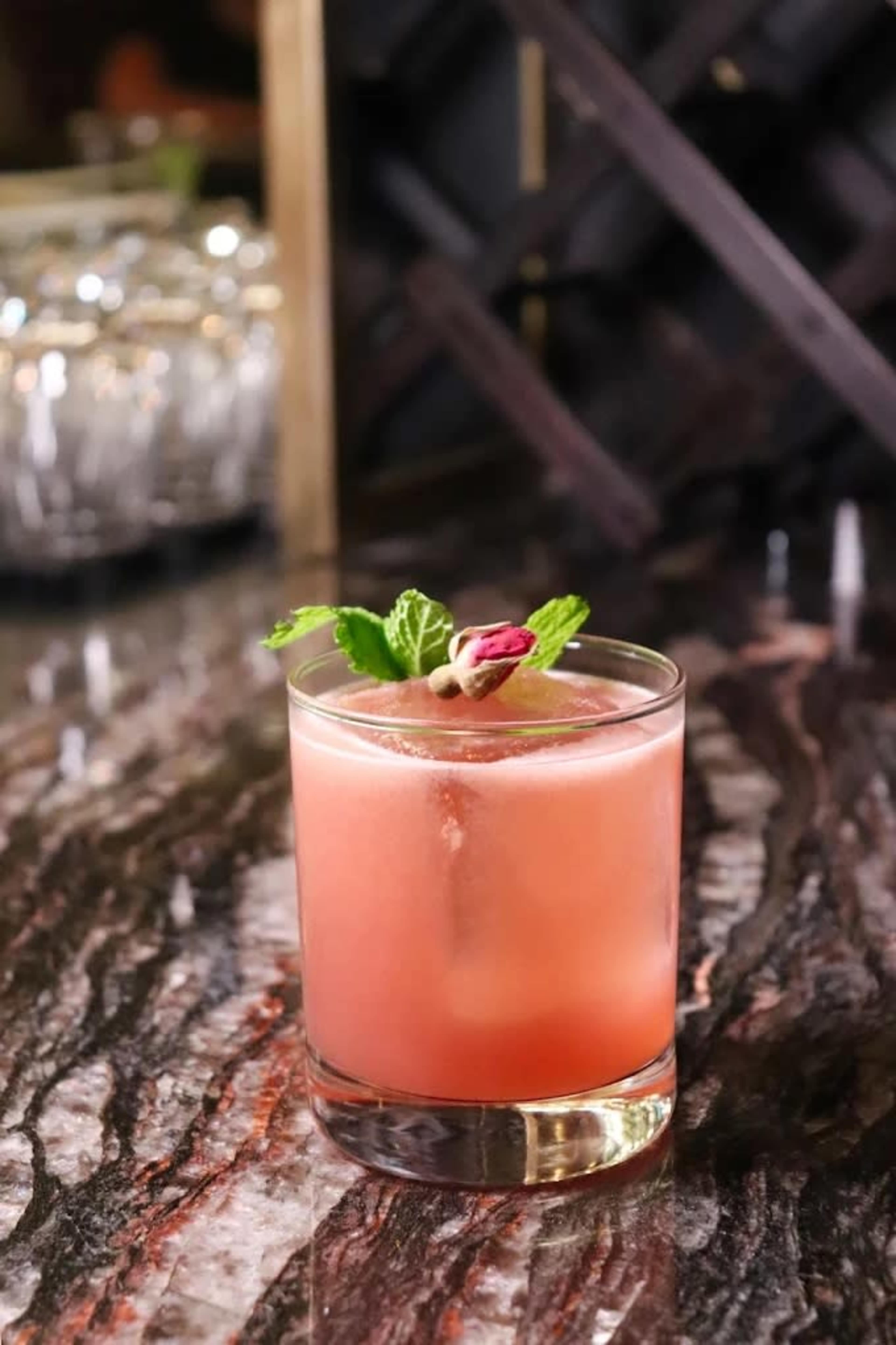 The image shows a cocktail served in a short glass, garnished with a mint leaf and a dried rosebud, placed on a marbled countertop.