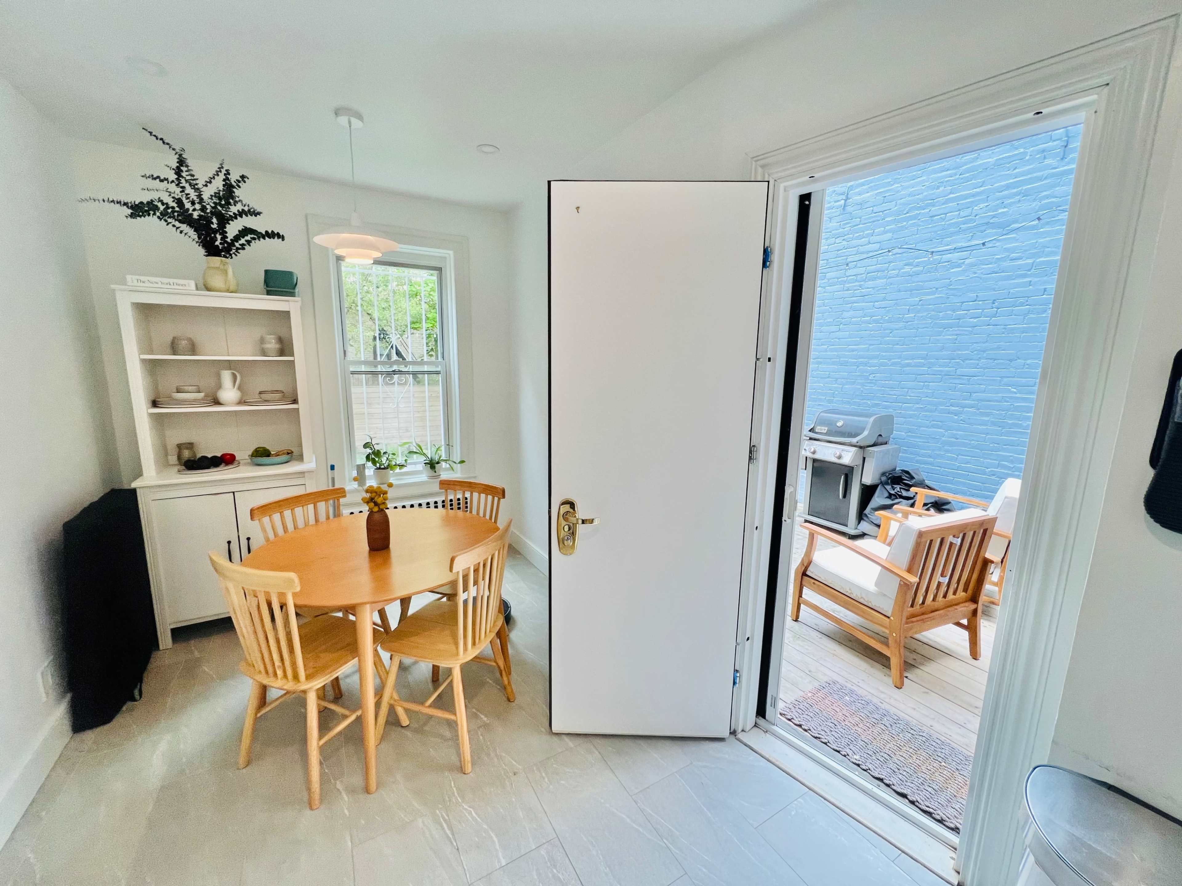 A bright kitchen area with a round wooden table surrounded by four chairs, a white cabinet, and a door leading to a small outdoor patio with seating and a grill.