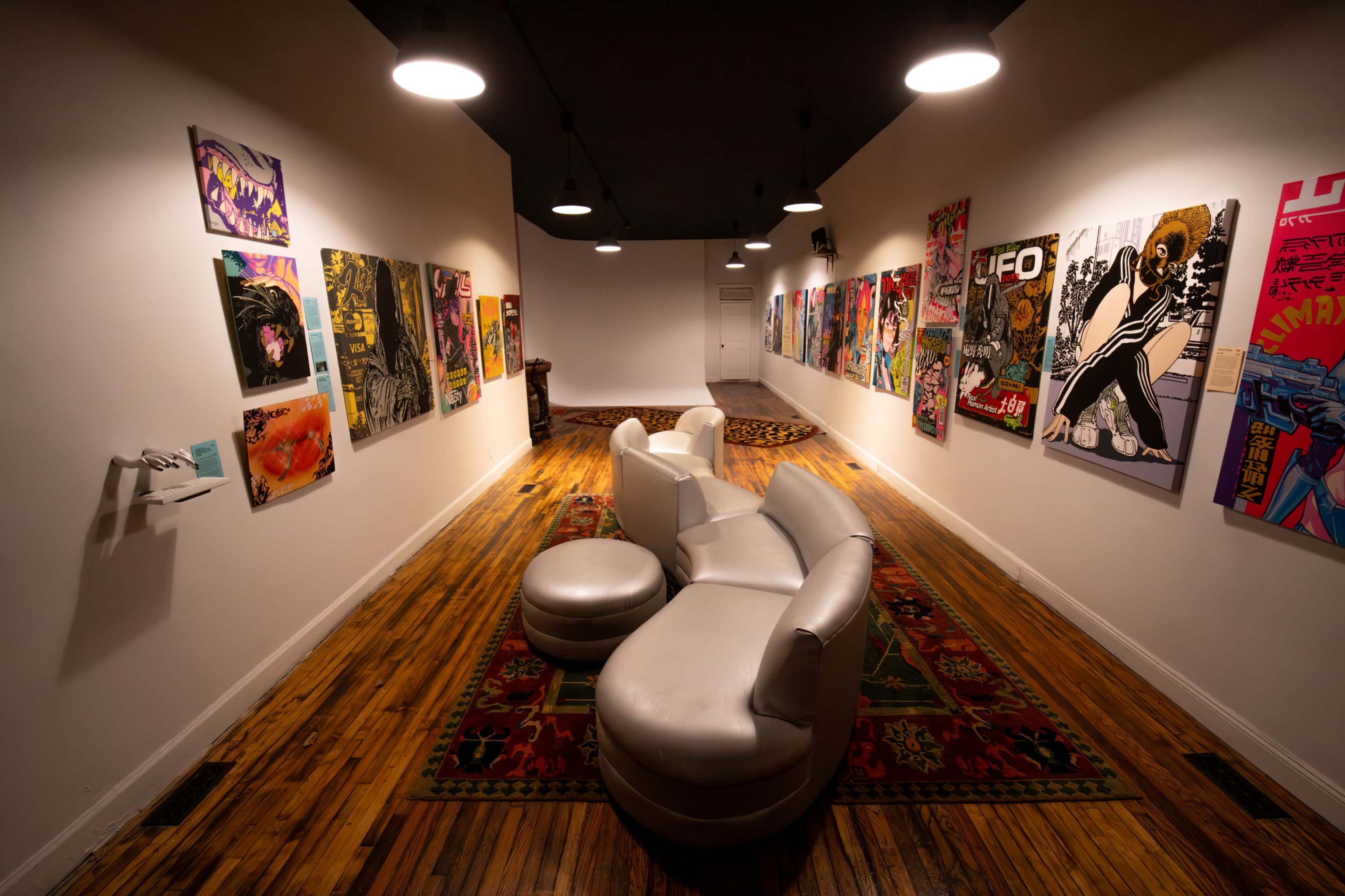 The image shows an art gallery with white walls showcasing colorful artwork, accompanied by a circular seating arrangement on a patterned rug.