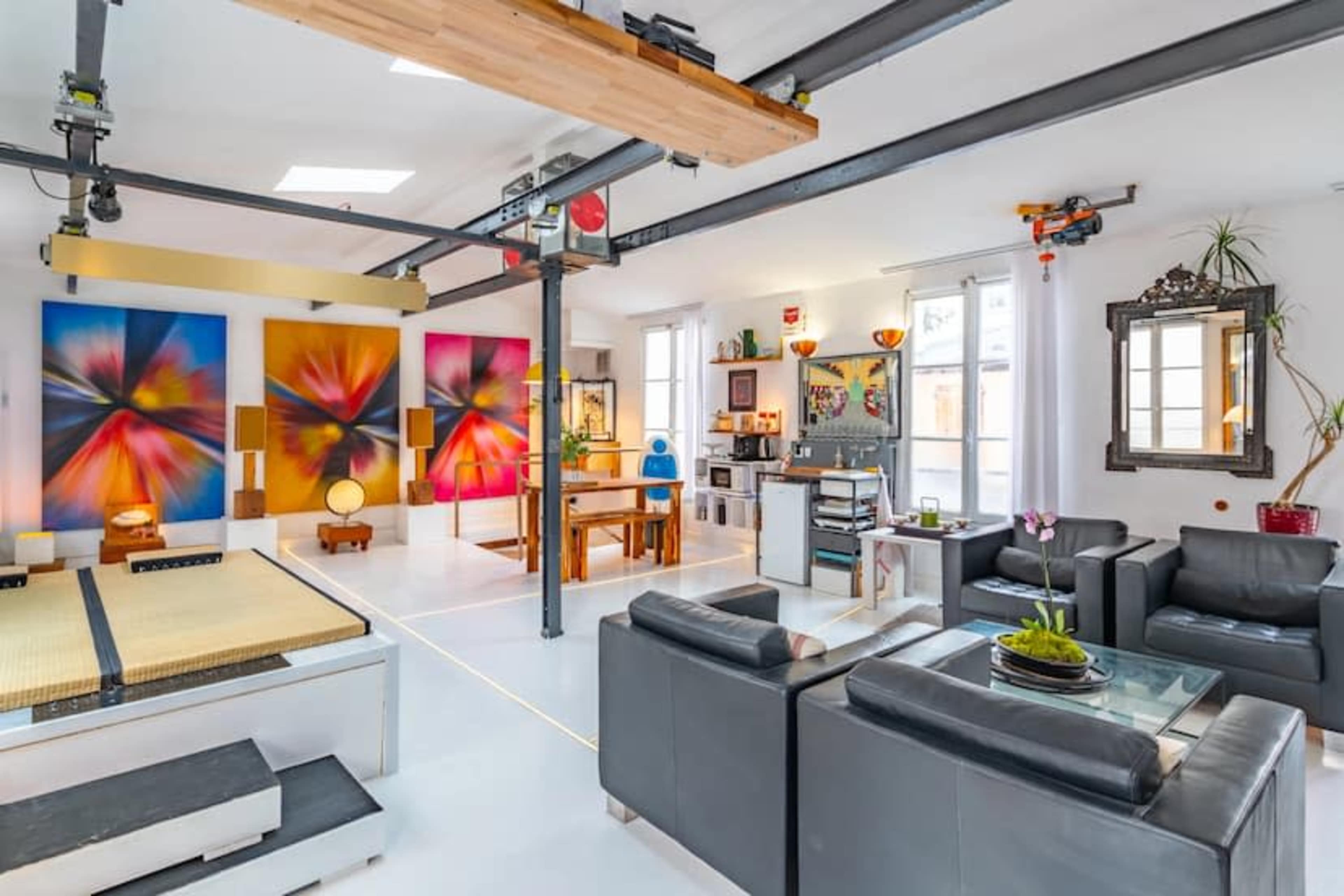 The image shows a spacious, modern living area with bright wall art, a dining table, and a seating arrangement featuring black leather sofas.