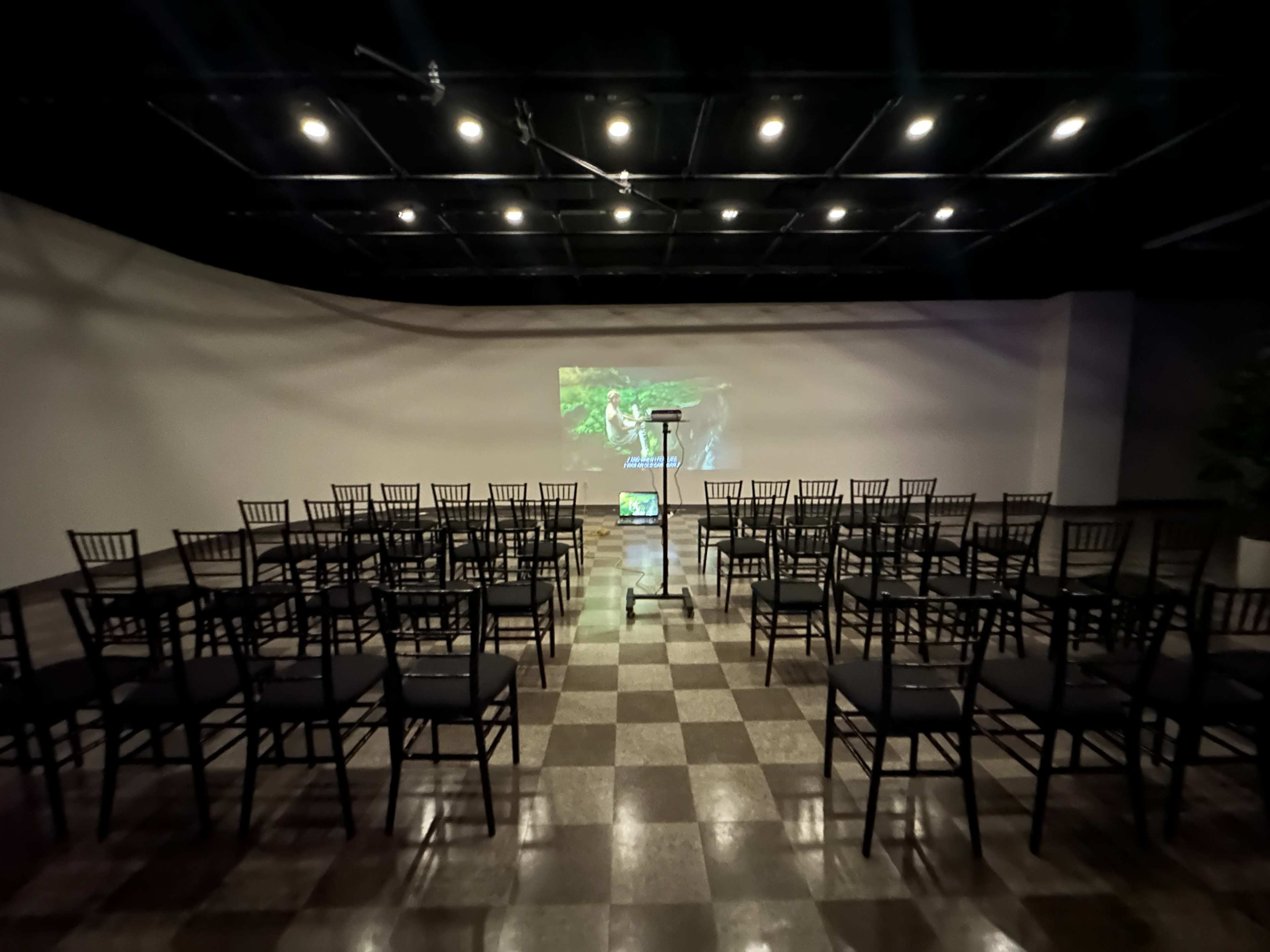 The image shows a large, empty room set up for an event, featuring rows of black chairs facing a podium and a projector screen displaying a green image on the wall.