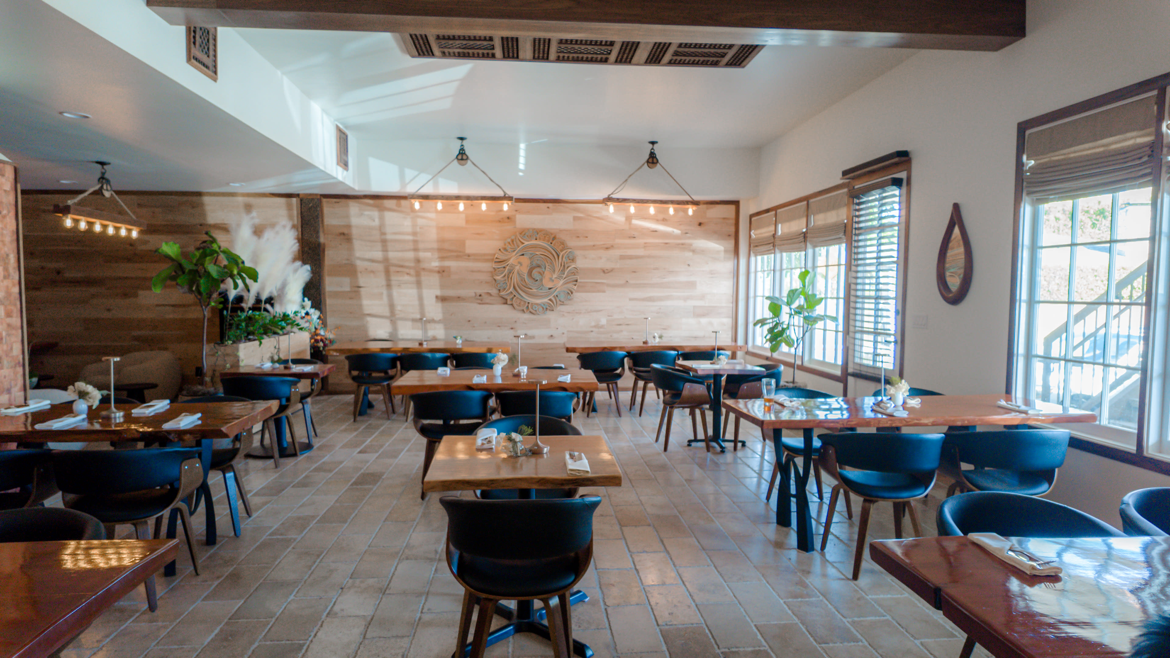The image shows a spacious restaurant with wooden tables and dark chairs, featuring a wooden wall with a decorative emblem and large windows allowing natural light.