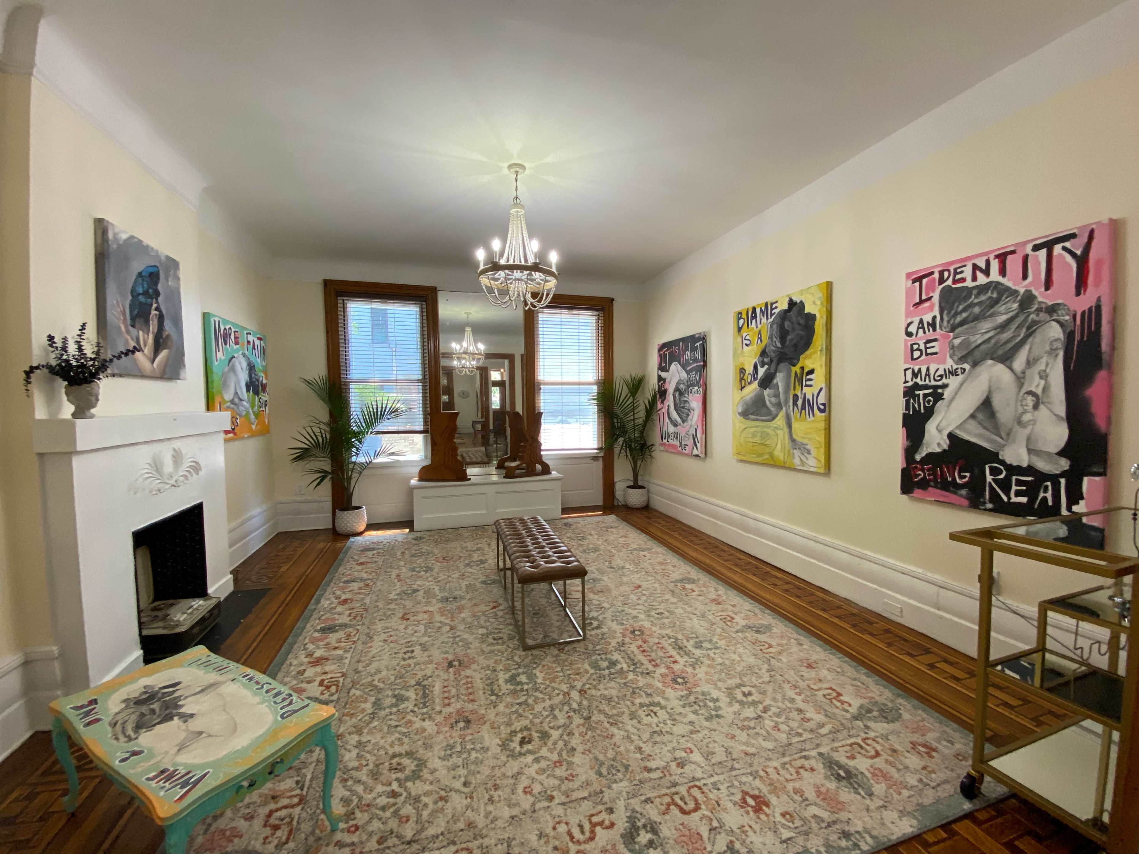 The room features a large carpet, a chandelier, and several colorful paintings on the walls, with a mirror and plants adding to the decor.