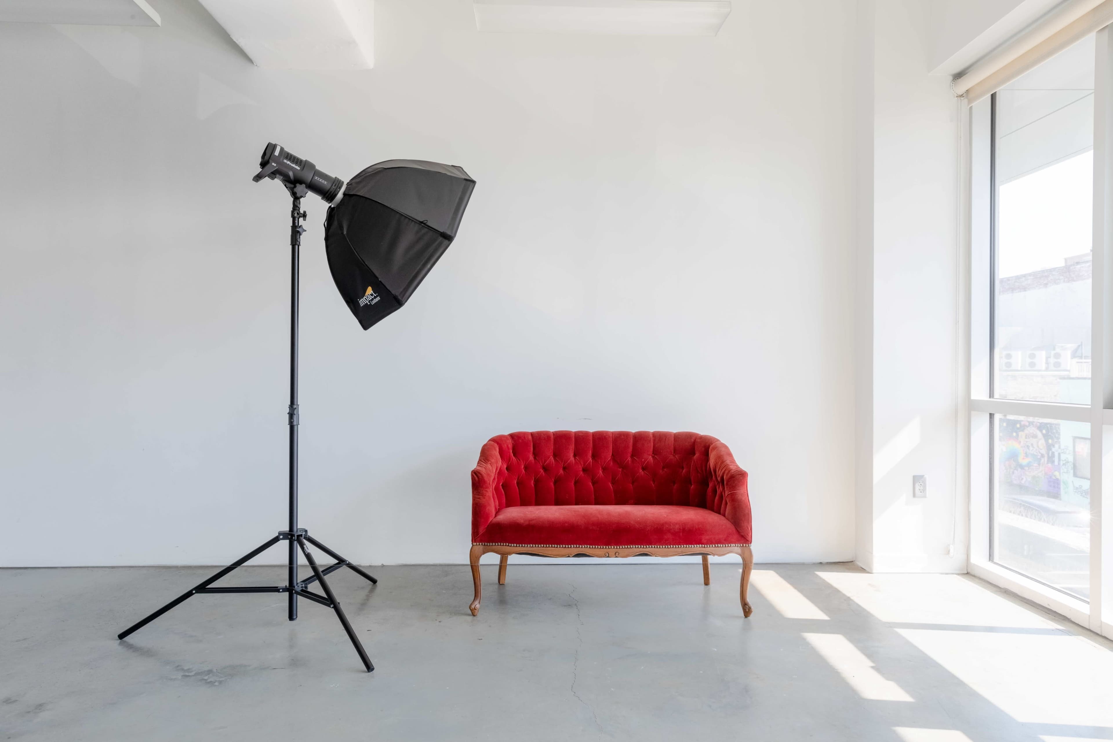 A red velvet sofa is positioned next to a large window, with a black photography umbrella reflector on a stand nearby.