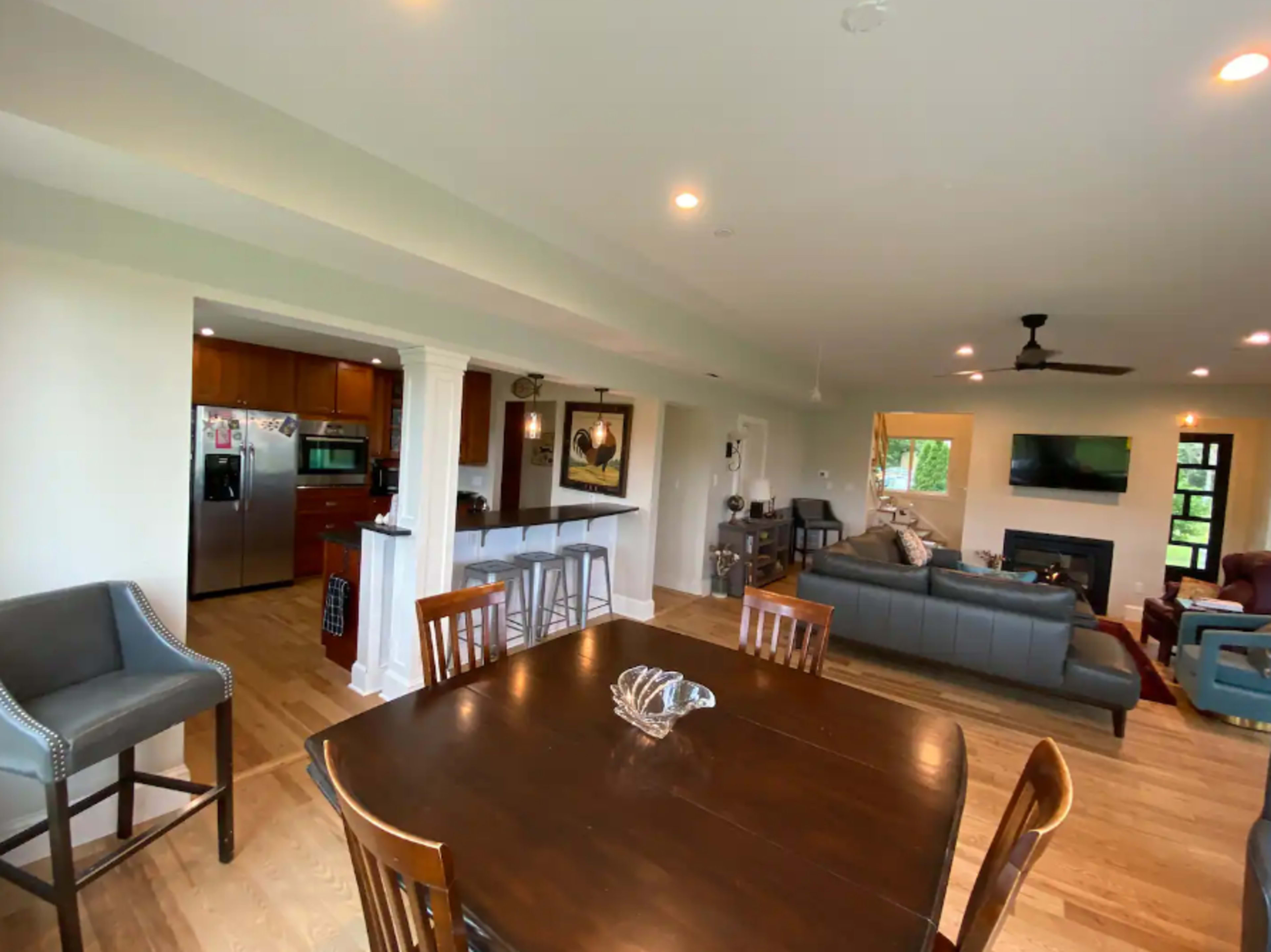 The image shows an open-concept living space featuring a dining area with a wooden table, a kitchen with stainless steel appliances, and a seating area with a sofa and television.