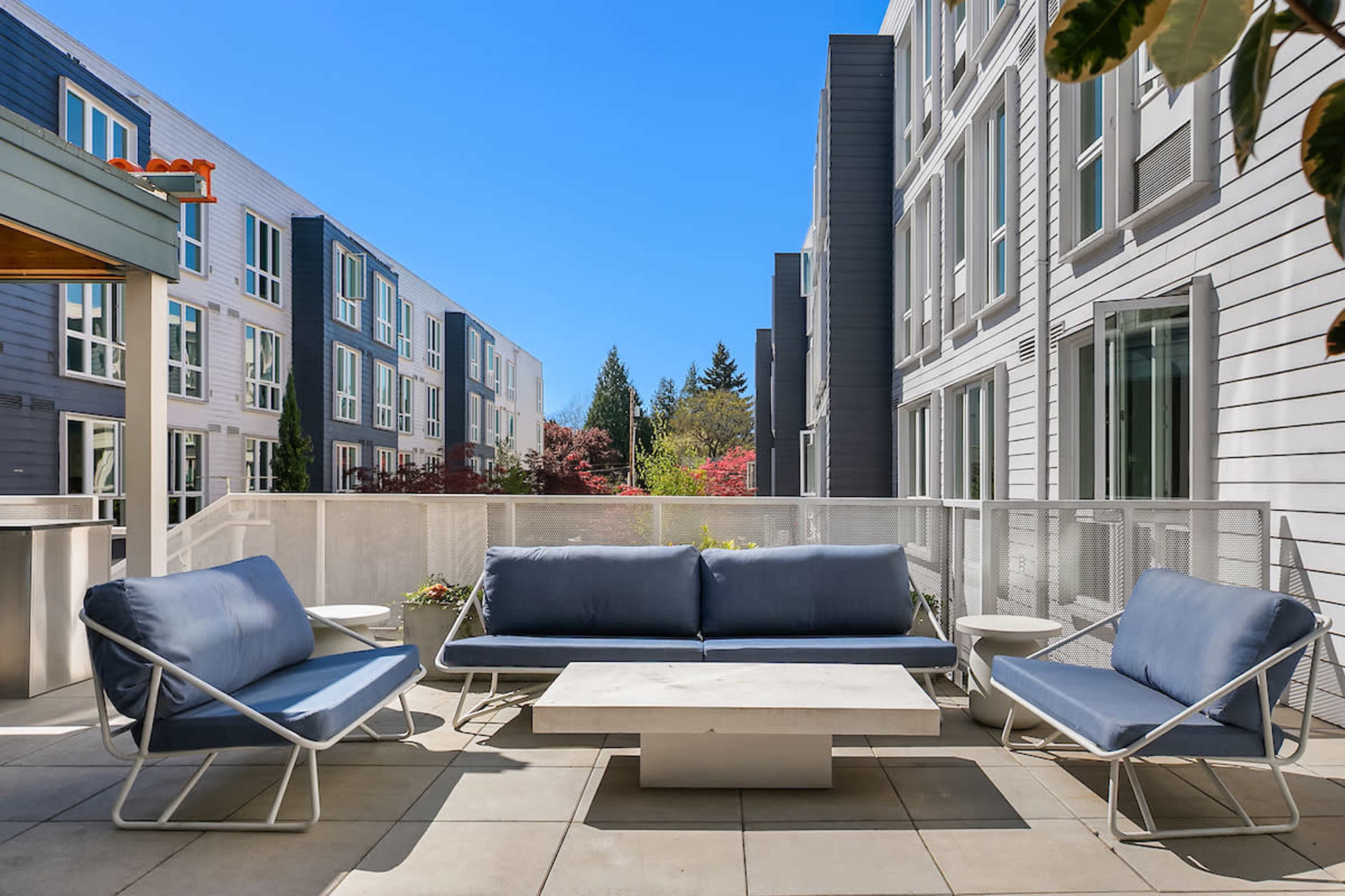 The image shows a modern outdoor lounge area with blue seating and a light-colored table, surrounded by contemporary apartment buildings.