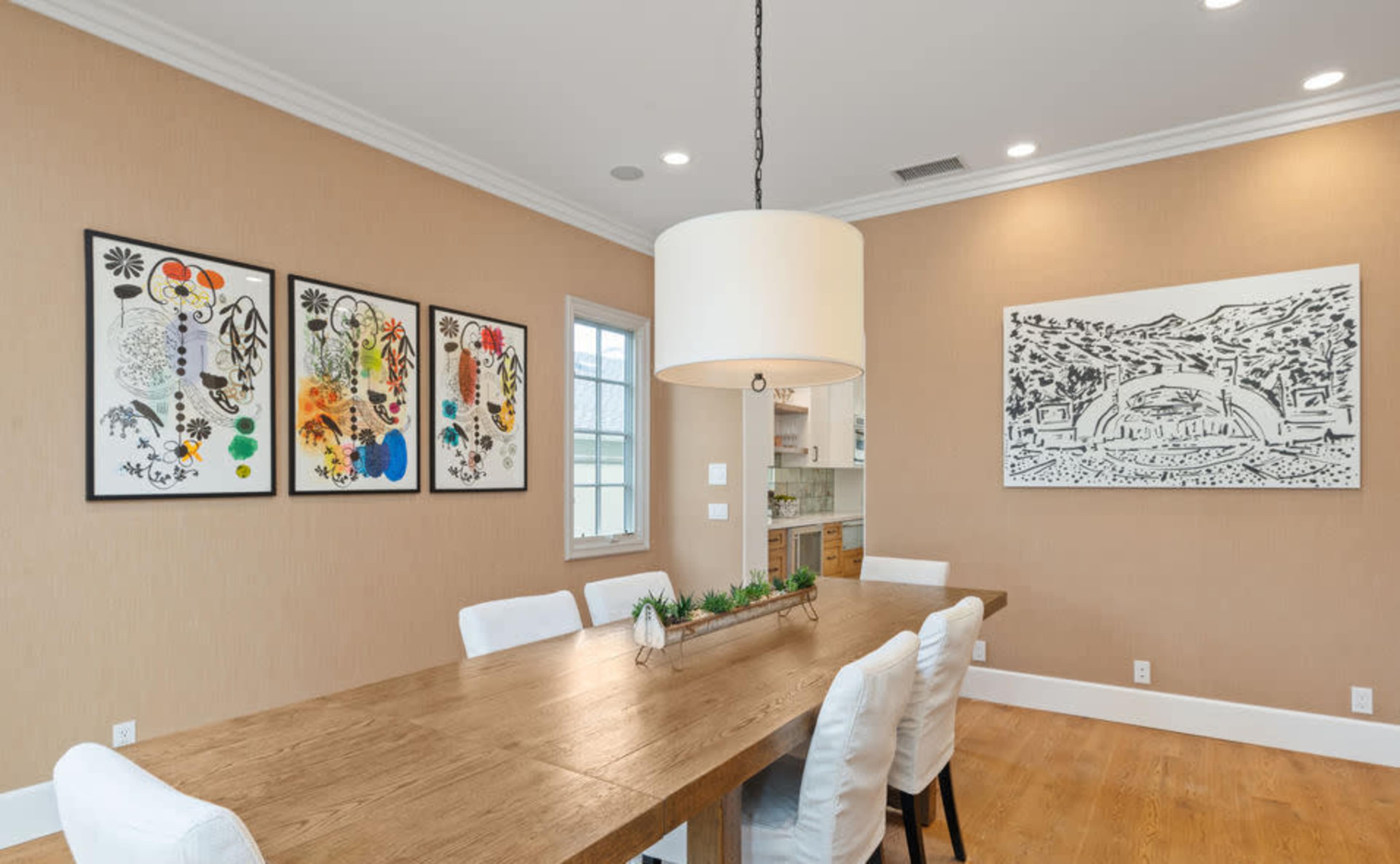 A dining room features a long wooden table surrounded by white chairs, with colorful artwork on the walls and a large pendant light above.