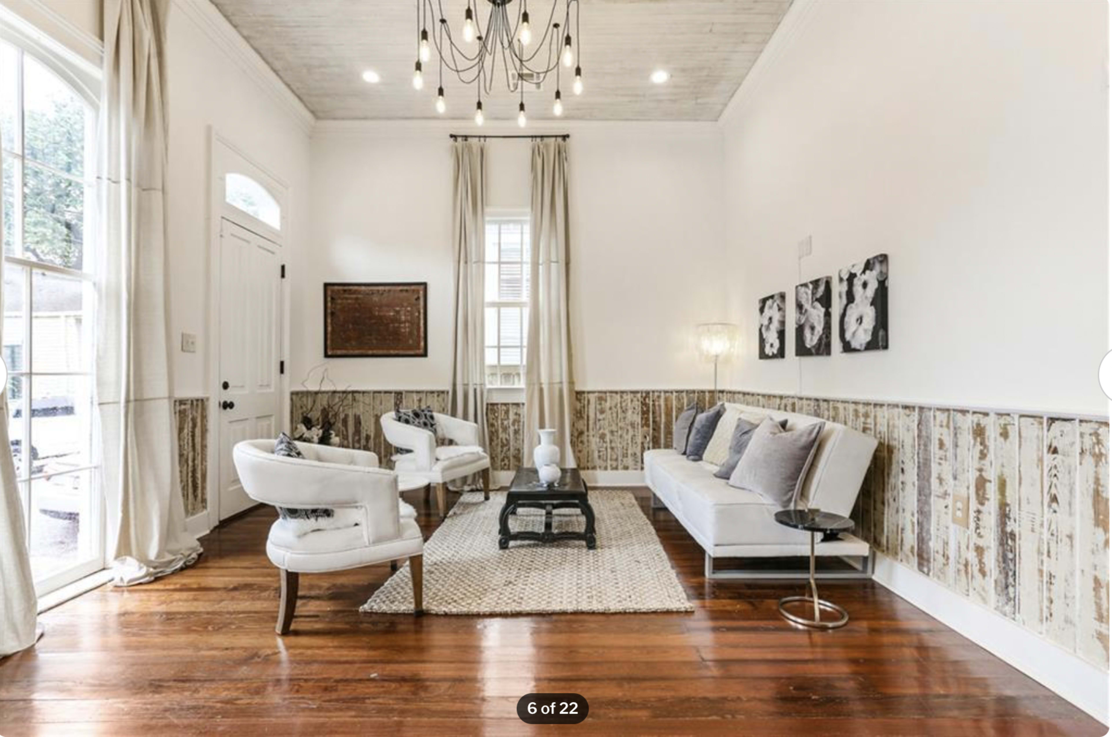 A bright and airy living room features a white sofa, two chairs, a decorative rug, and wooden walls with artwork.