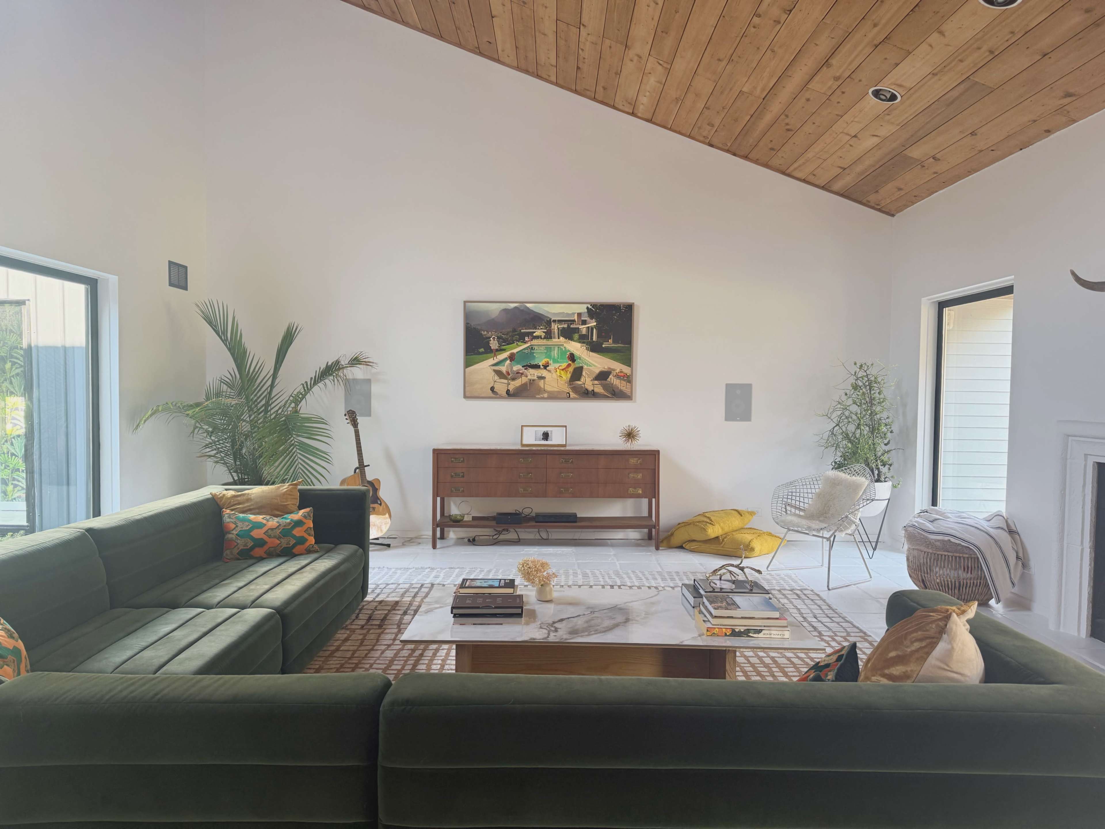 A modern living room features a green sectional sofa, a wooden media console, and a photograph on the wall, with plants and decorative pillows arranged throughout the space.