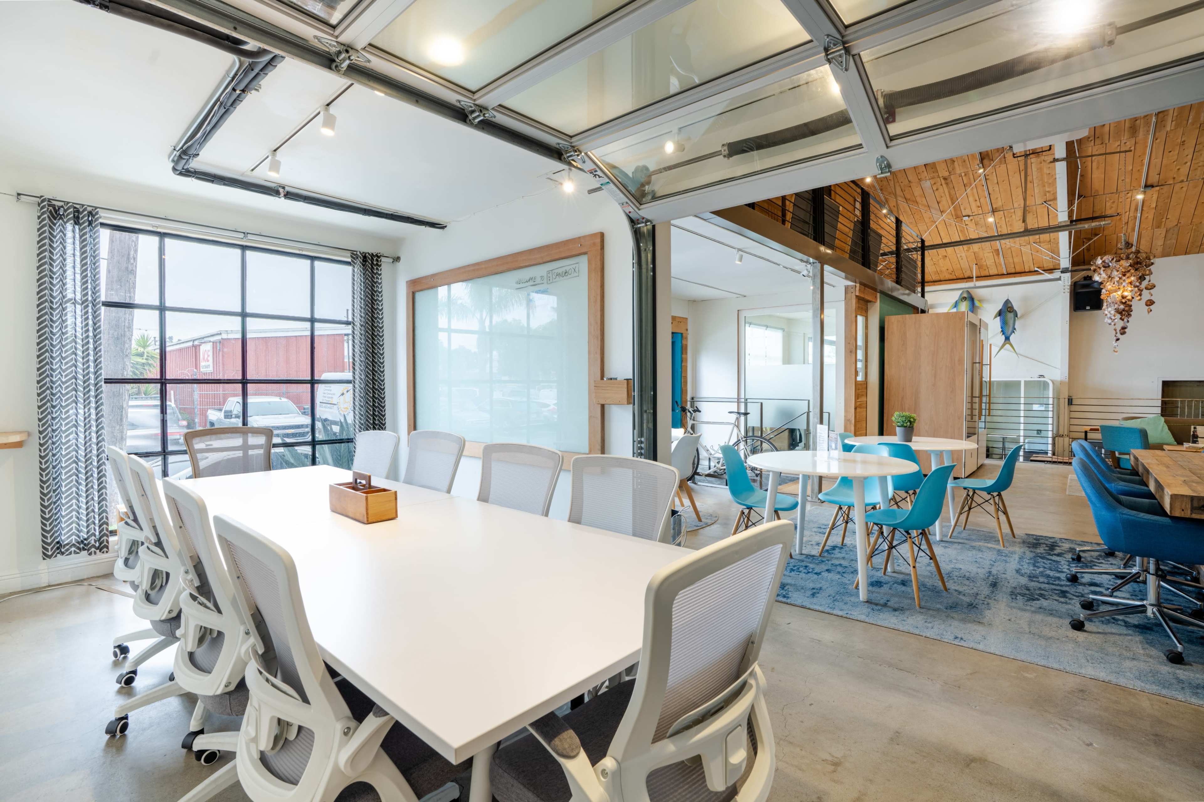 The image shows a modern office space featuring a large meeting table surrounded by white chairs, with glass walls and an open layout that includes a blend of workstations and casual seating areas.