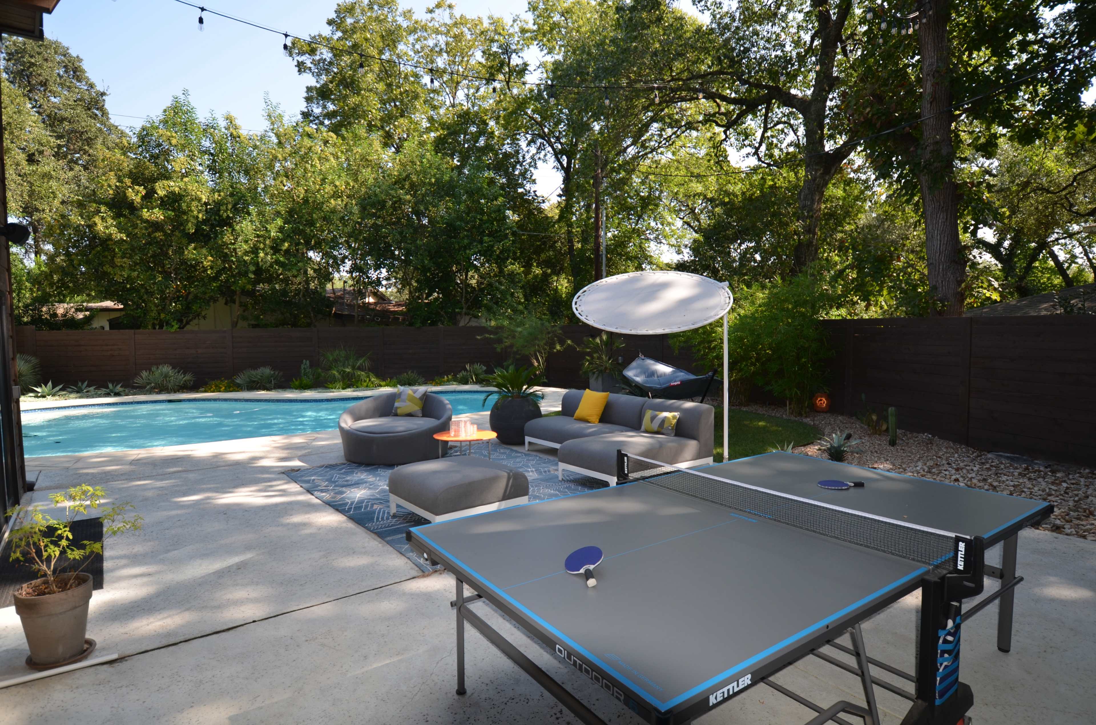 The image shows a backyard with a ping pong table in the foreground, a swimming pool in the background, and outdoor seating arrangements beside the pool.