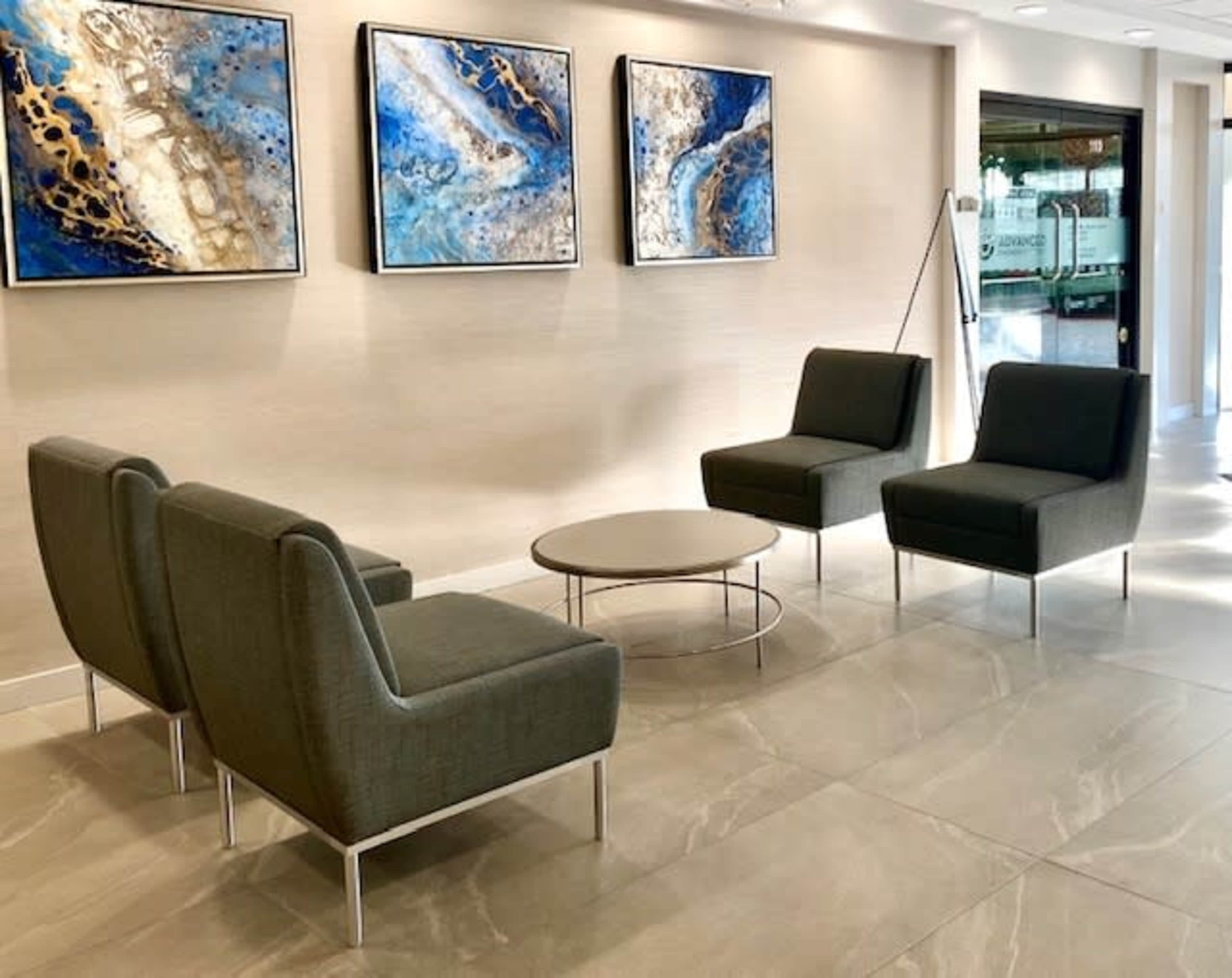 A modern waiting area with two green chairs, a round coffee table, and three abstract paintings on the wall.