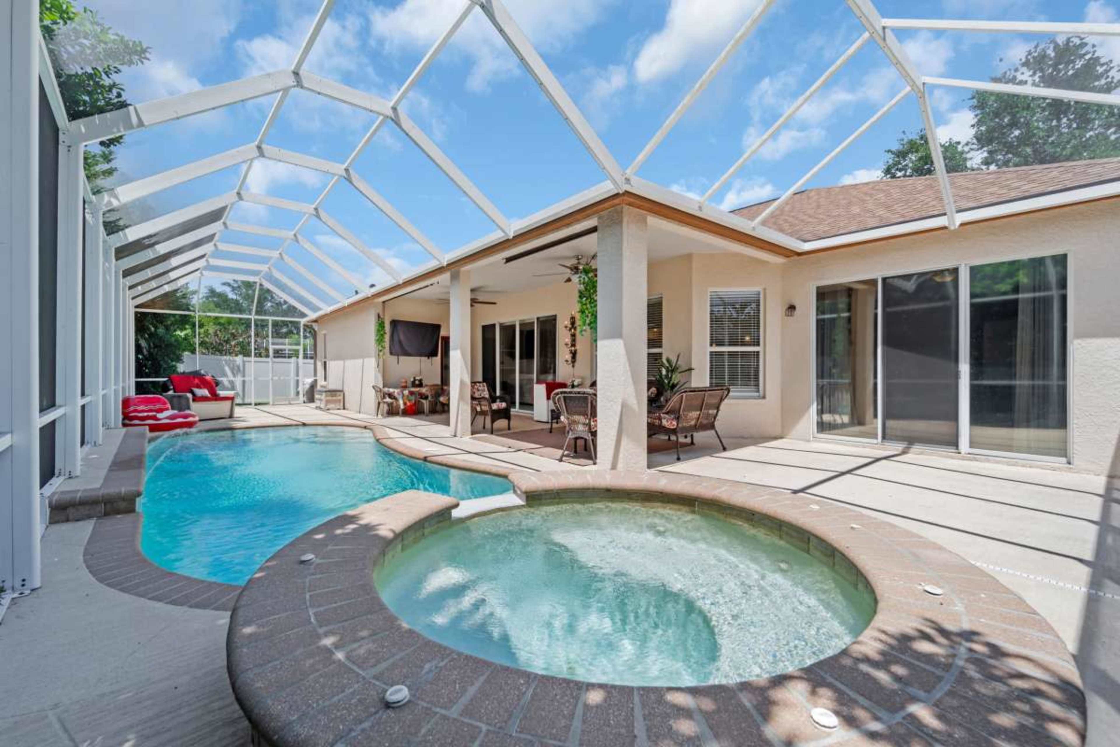 The image shows a pool area enclosed by a screened roof, featuring a swimming pool and a jacuzzi beside a house with outdoor seating.