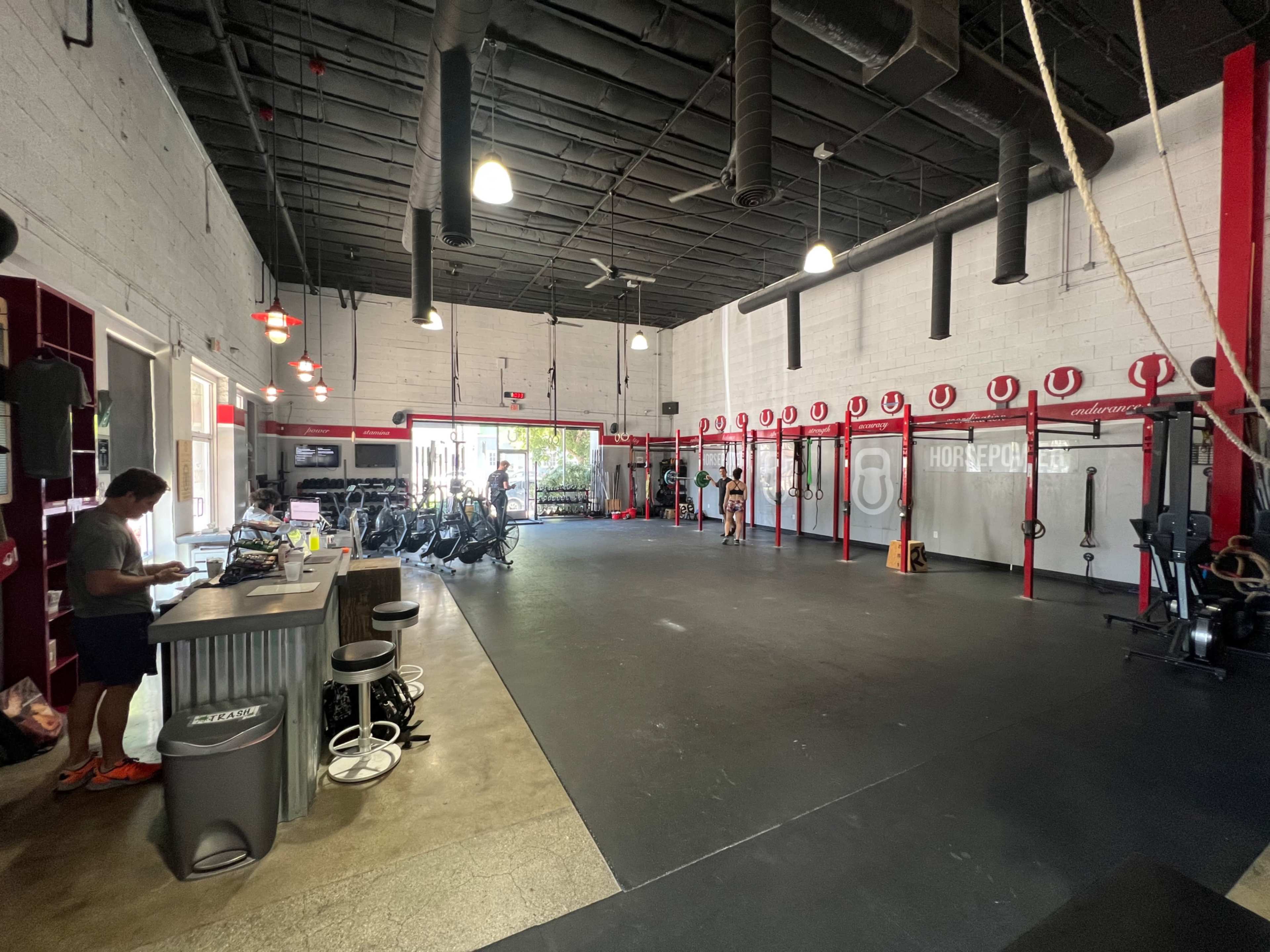 An interior view of a gym with exercise equipment arranged against the walls and a few people engaged in workouts.