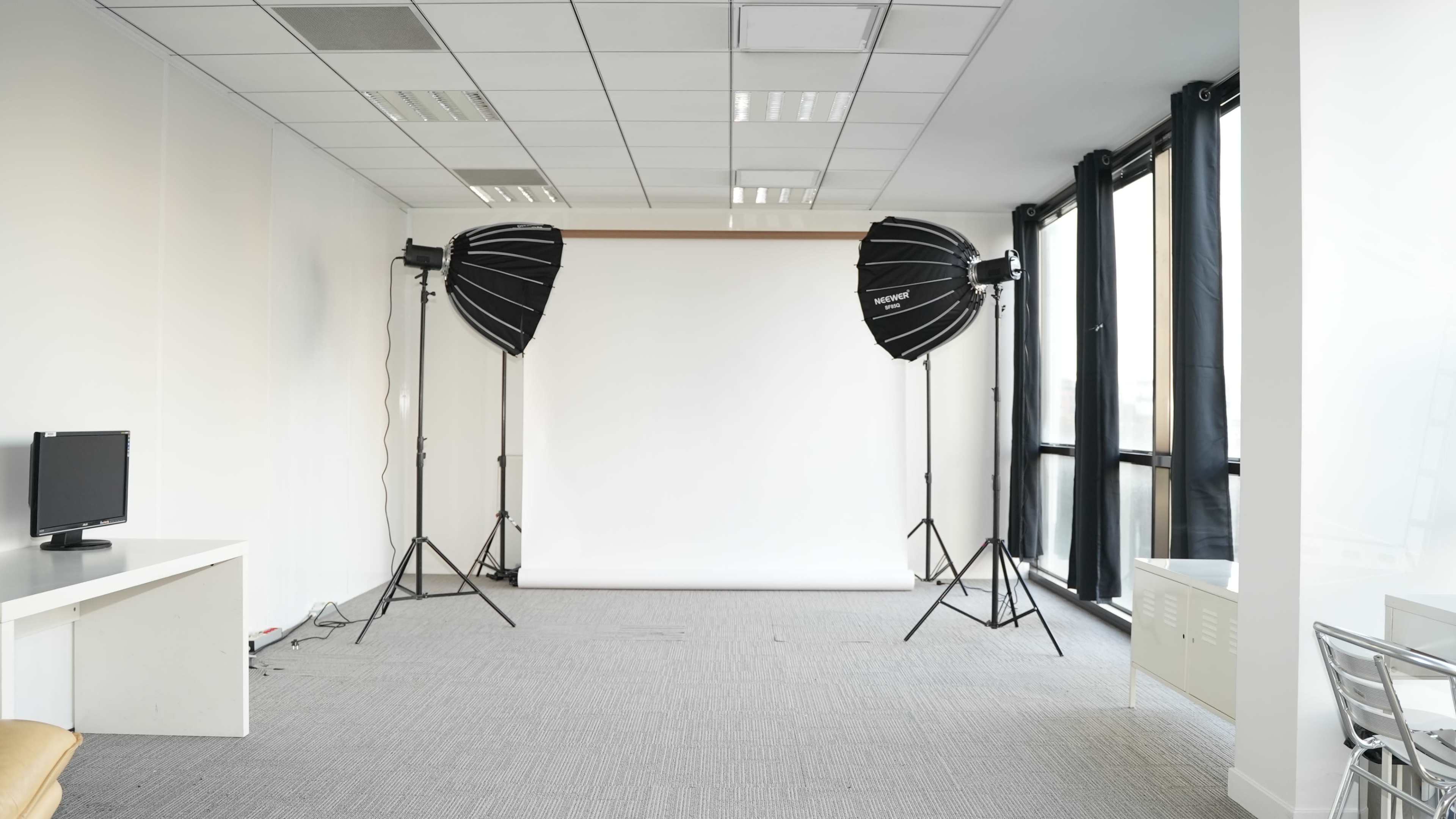 The image shows a spacious studio with a white backdrop, two softbox lights on tripods, and large windows on one side.