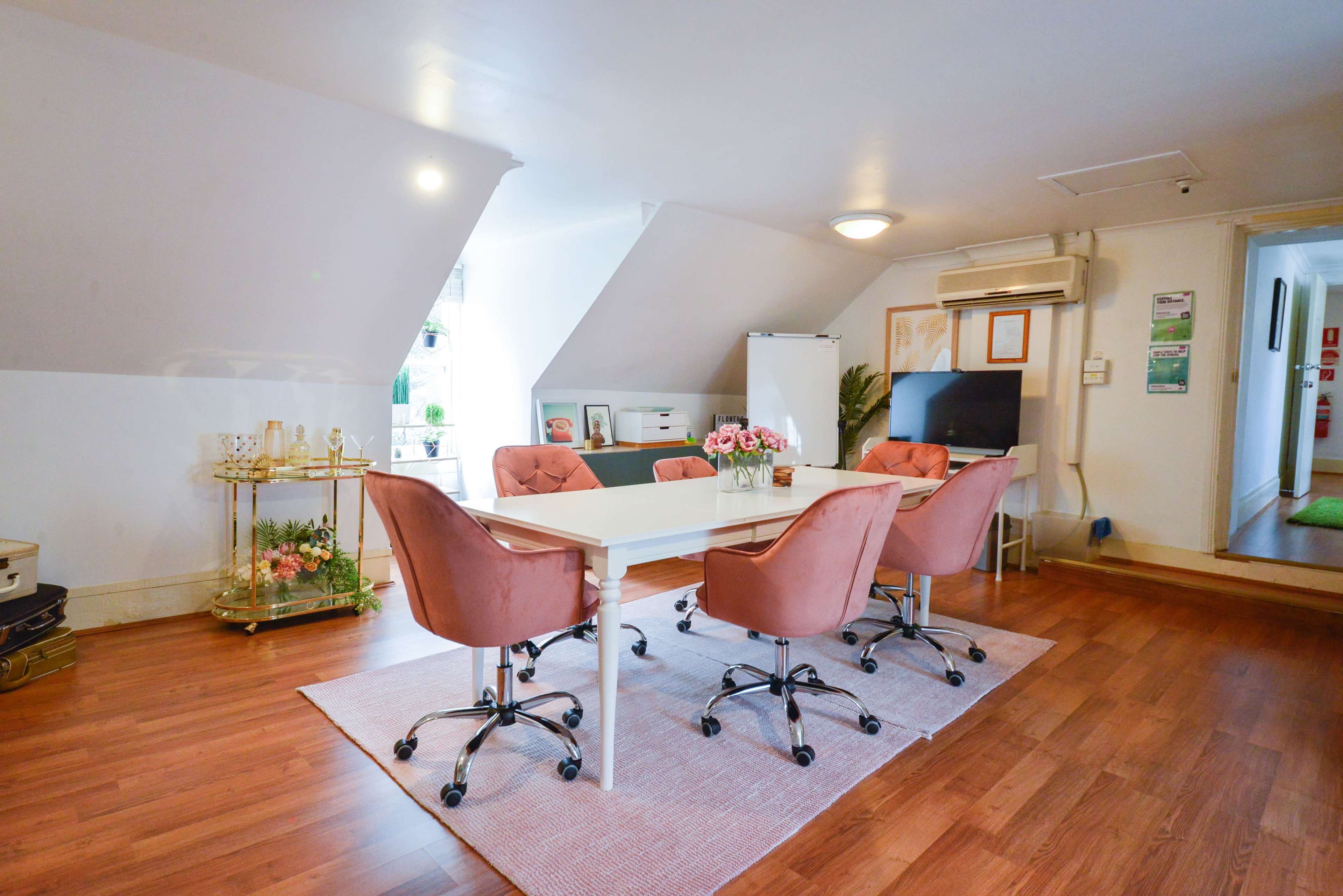 A modern meeting room with a white table surrounded by pink chairs, a notice board, and decorative plants.
