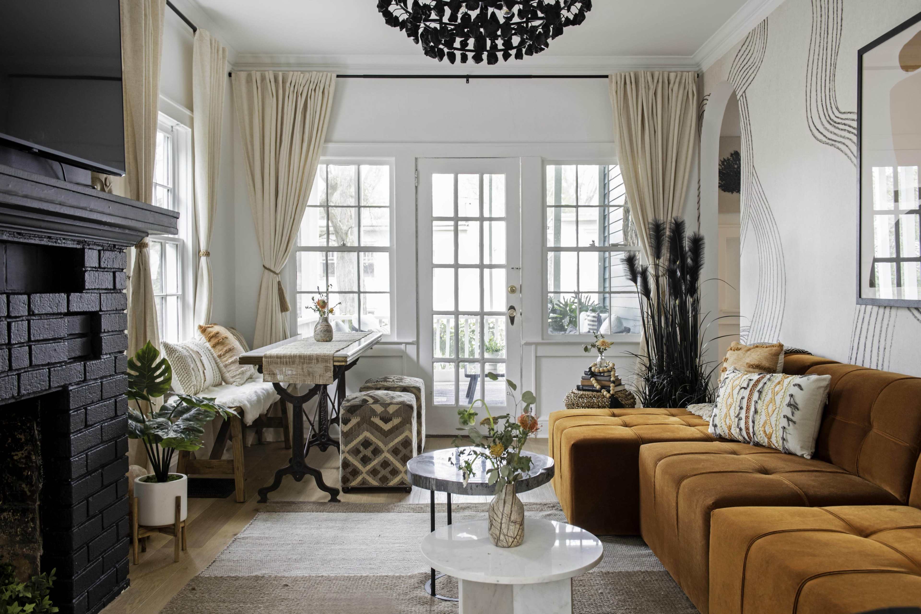 The image shows a modern living room with a brown sectional sofa, a white coffee table, a decorative black chandelier, and large windows that provide natural light.